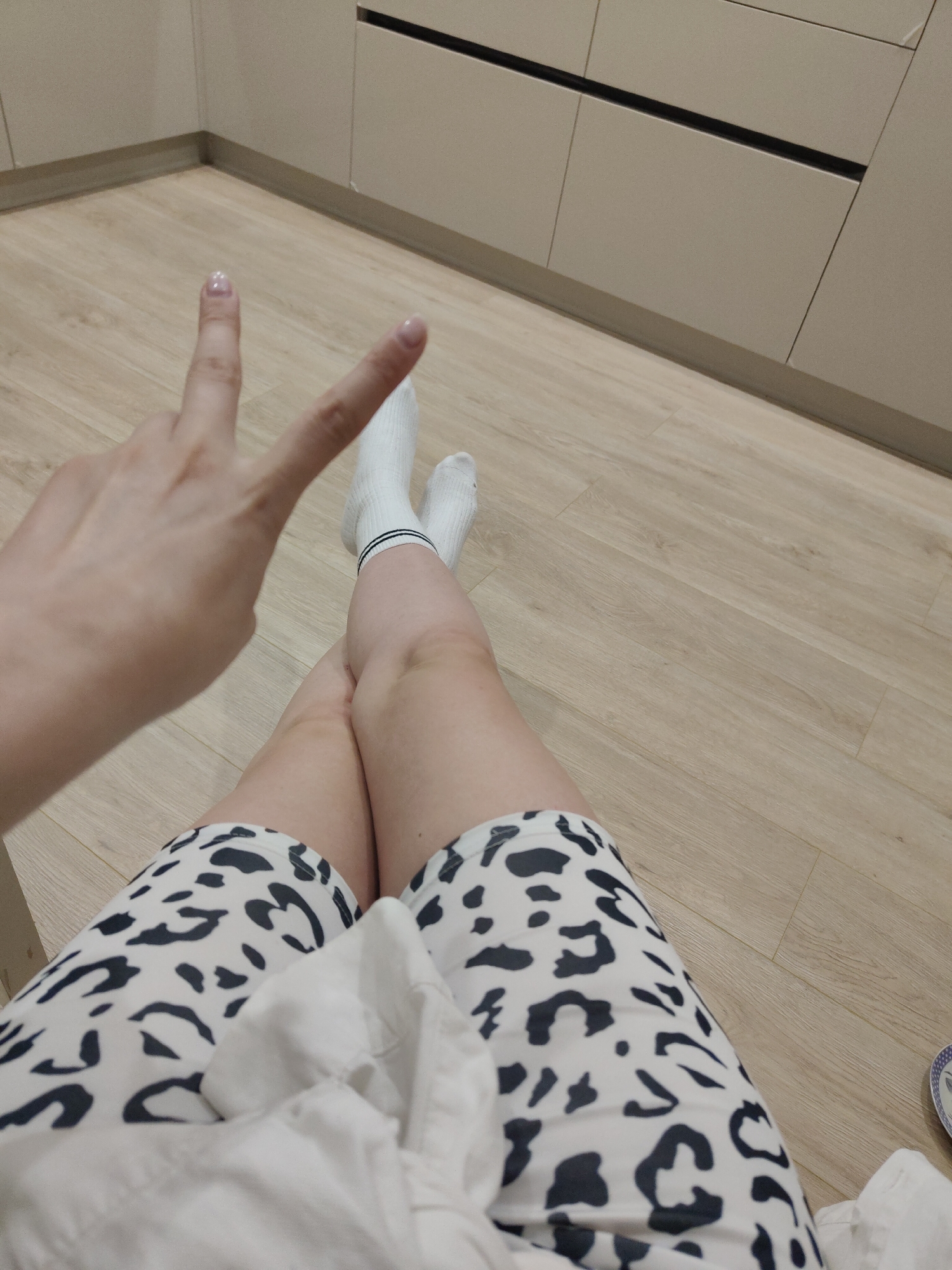 The photograph shows a person sitting on a wooden floor. The person is wearing white socks with black stripes and a pair of white shorts with a black animal print. They are making a peace sign with their hand. The background includes kitchen cabinets. The location is Unknown, Unknown.