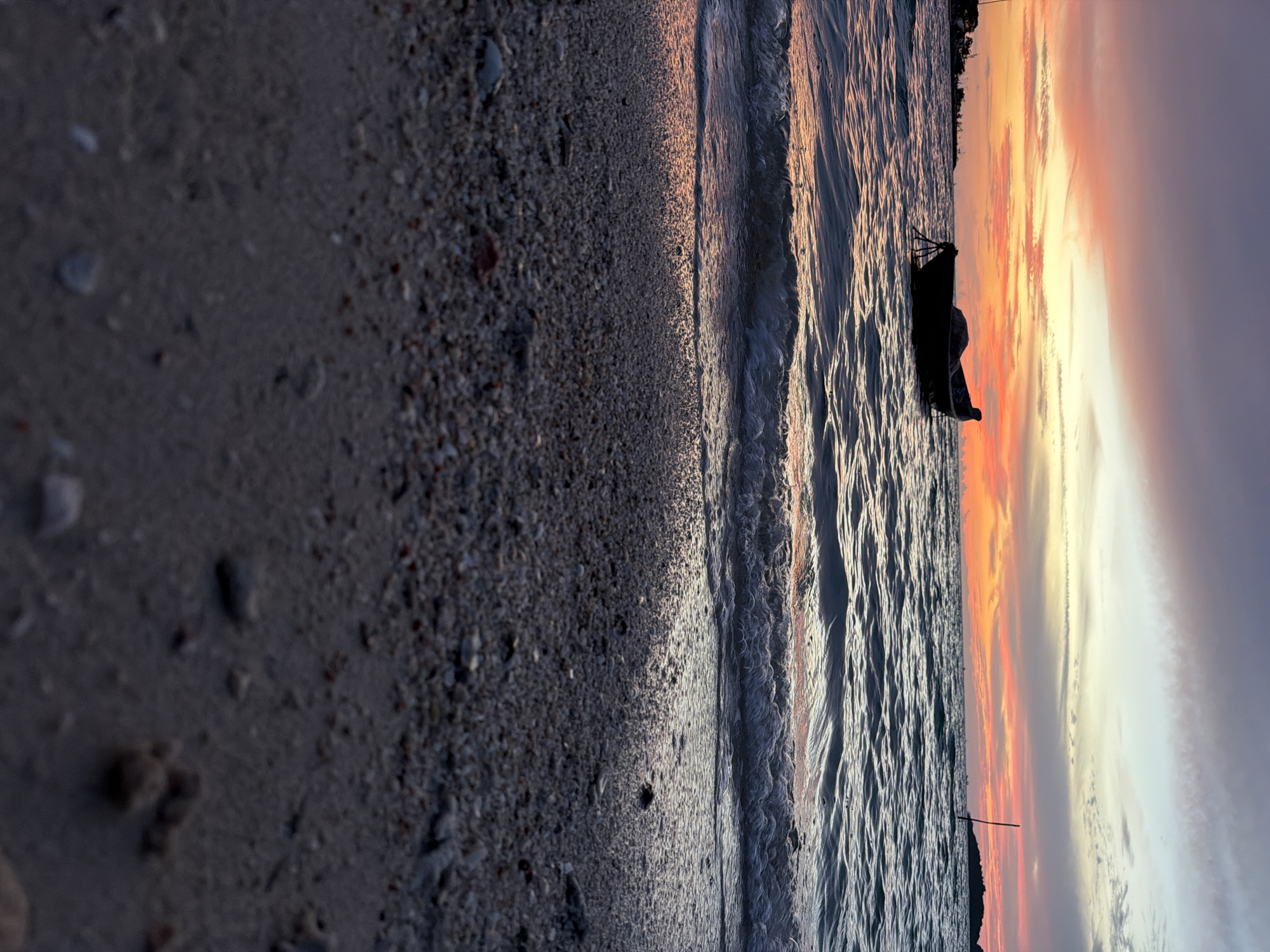 A small, dark-colored boat is anchored in the water near a beach, as gentle waves lap the shore. The sky above is a vibrant canvas of sunset colors, with hues of orange, pink, and purple blending together. The foreground is dominated by the texture of the sandy beach, dotted with small pebbles and shells. In the distance, the outline of land and a few indistinct structures are visible against the colorful sky. The scene evokes a sense of tranquility and the peaceful end of a day. This image likely depicts a scene in the Ko Samui District of Thailand, known for its beautiful beaches and stunning sunsets.