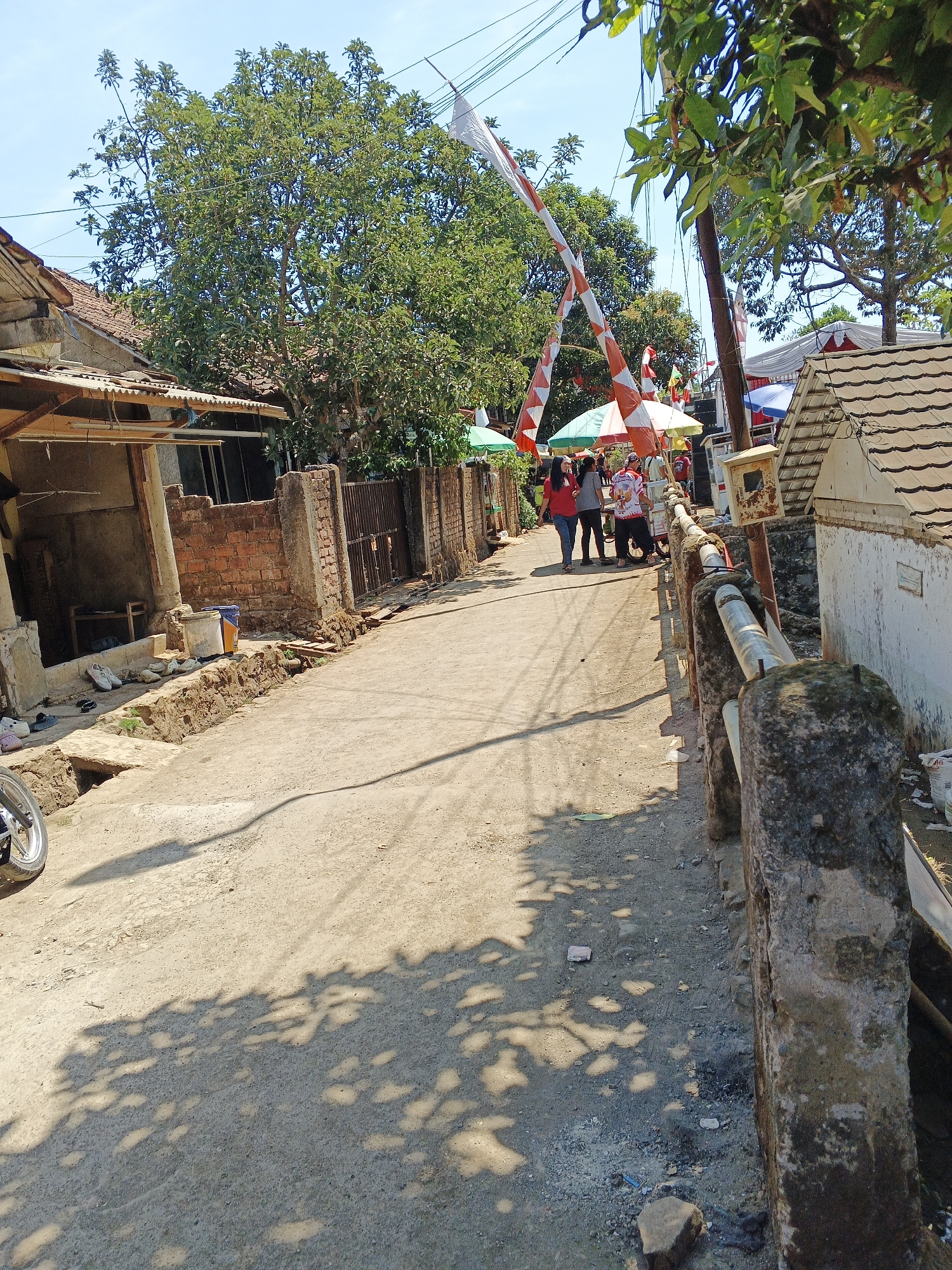 In Sukaharja, Indonesia, on a sunny day, a narrow dirt road runs through a residential area, lined with simple brick and concrete buildings. Several people are gathered near the center of the frame, some standing under umbrellas and decorated with red and white flags, suggesting a local celebration or event. Two women in the foreground walk along the road, dressed casually. On the left side of the road, there's a structure with exposed brick walls and a corrugated metal roof, with shoes and a bucket placed near its base. A motorcycle wheel is visible on the far left. A concrete fence with a metal railing runs along the right side of the road, separating it from a white building with a tiled roof. Trees with green foliage are abundant, casting dappled shadows across the road, and utility poles with wires are present in the background. The overall impression is of a modest, everyday scene in a rural Indonesian community.