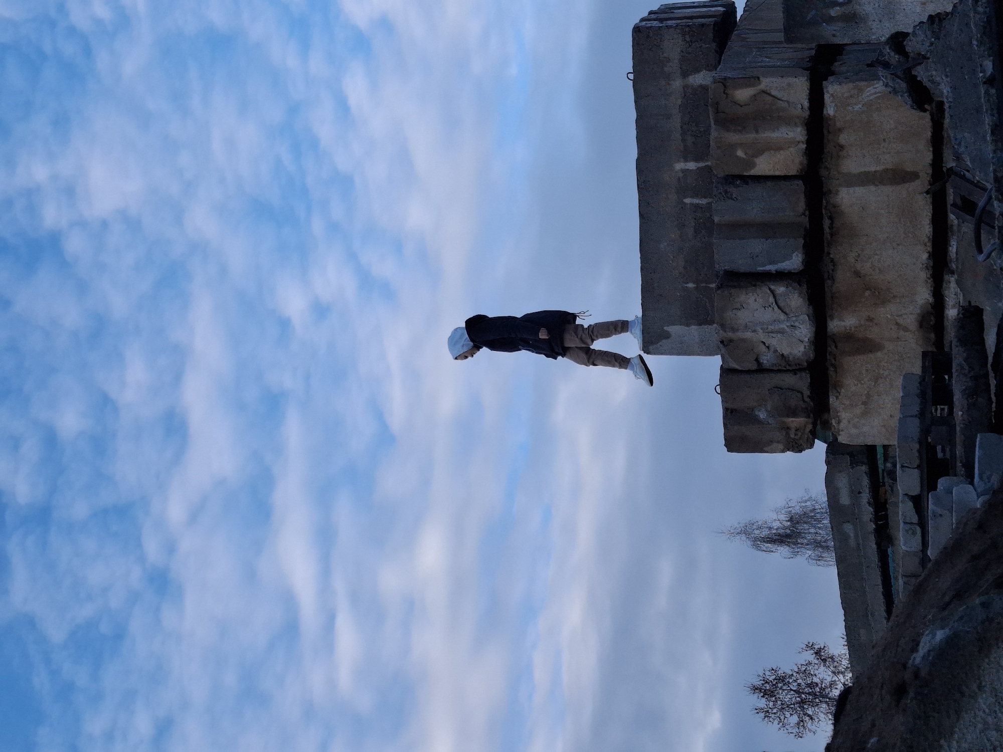 A full shot depicts an individual standing on the edge of a concrete structure against a sky of patchy white clouds on a blue background. The individual wears a dark jacket, tan pants, white shoes, and a white beanie. The concrete structure appears to be a crumbling building or ruin, composed of gray and weathered blocks. A bare tree branches out near the structure’s base. The scene is in Chaikovsky, Russia.