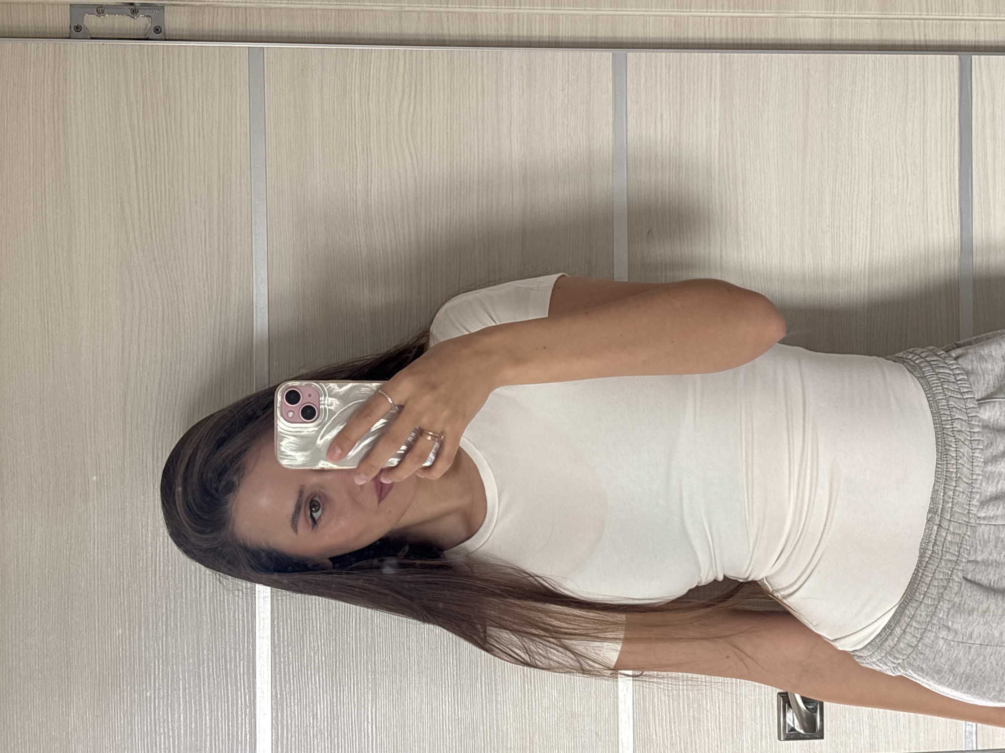 A woman is taking a selfie in front of a wooden-paneled wall, likely a door. She is holding a smartphone in her right hand, with a clear case. She has long, brown hair and is wearing a white T-shirt and gray sweatpants. The location is Krasnoyarsk, Russia. The photograph seems to be taken indoors.