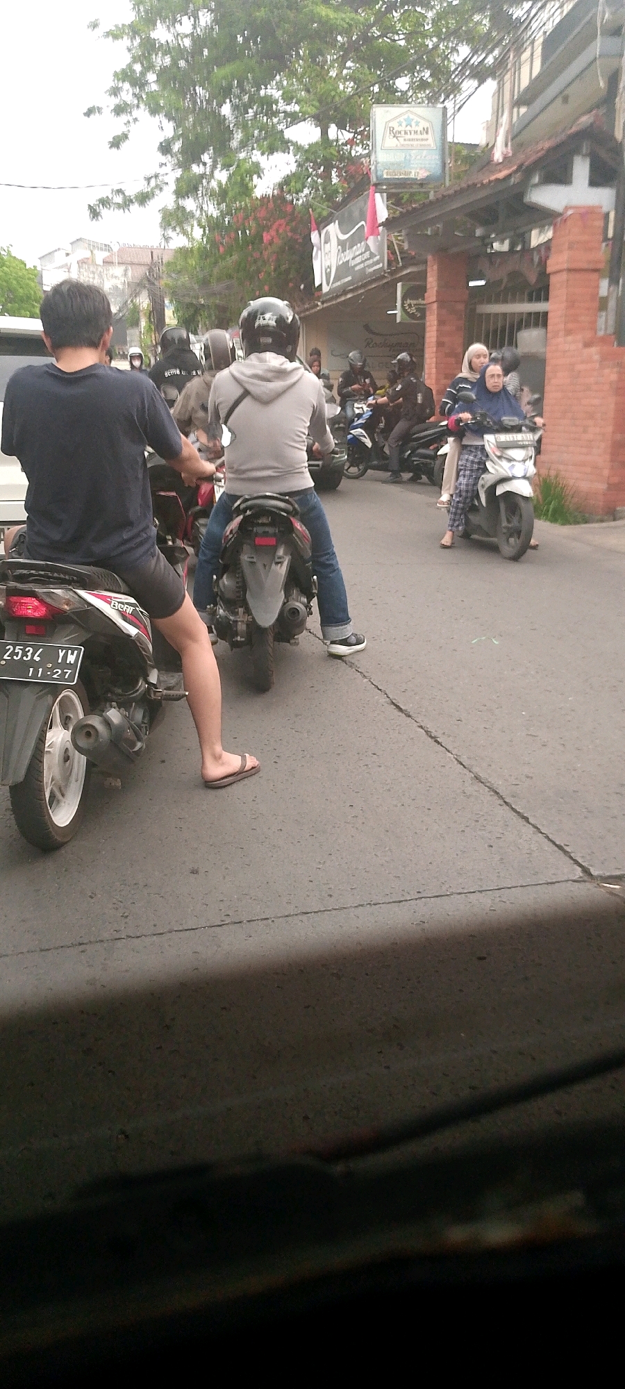 The image captures a street scene in Bandung City, Indonesia, likely during the daytime given the lighting. In the foreground, a person is seated on a black and white motorcycle, facing away from the camera. They are wearing a dark t-shirt and shorts, with one leg extended, wearing a flip-flop. The motorcycle has a license plate reading "2534 YW 11-27". To the right of this motorcycle, another person is seated on a similar motorcycle, wearing a grey hoodie and blue jeans. This motorcycle is also facing away from the camera. Further down the street, a white motorcycle is visible with two occupants. The person in front is wearing a headscarf and a blue top, while the person behind them is wearing a helmet. Other motorcycles and people are present in the background, suggesting a moderately busy street. Buildings line the side of the street, with signage visible including what appears to be a "ROCKYMAN" sign. Trees with green foliage are also present, indicating an urban environment with some greenery. The road surface is asphalt with visible cracks and markings. The overall impression is of everyday street traffic in a city.