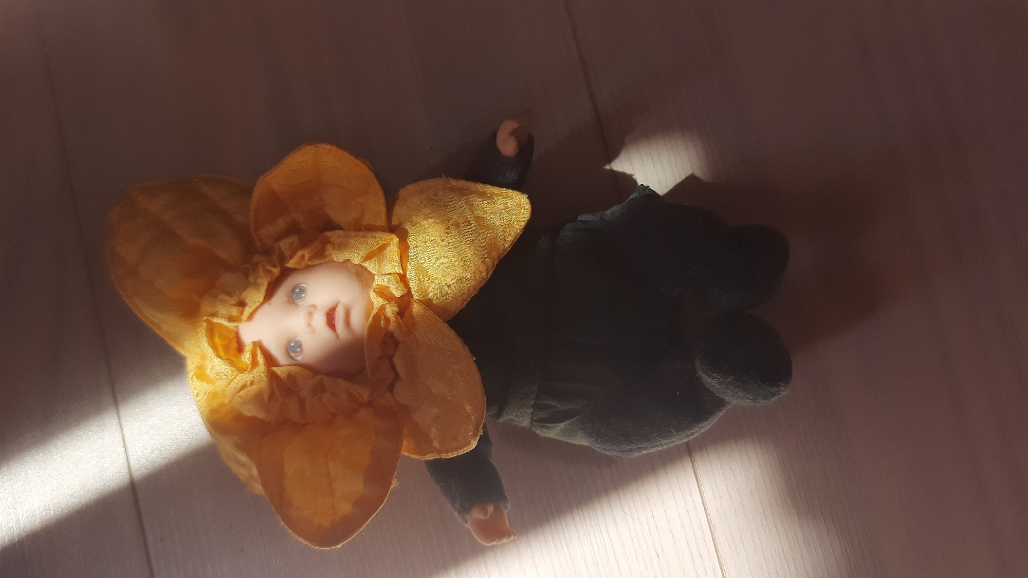 A baby doll wearing a sunflower costume is lying on a light-colored wooden floor, illuminated by diagonal rays of sunlight. The doll has blue eyes and its mouth is slightly open, giving it an surprised expression. The costume consists of yellow fabric petals forming a large flower head around the doll's face and a dark green body with black fabric for the legs. The doll is positioned with its head slightly to the right, and its body extends downwards and to the left. The lighting creates distinct shadows on the floor. The scene appears to be indoors, possibly in a residential setting in Desnogorsk, Russia.