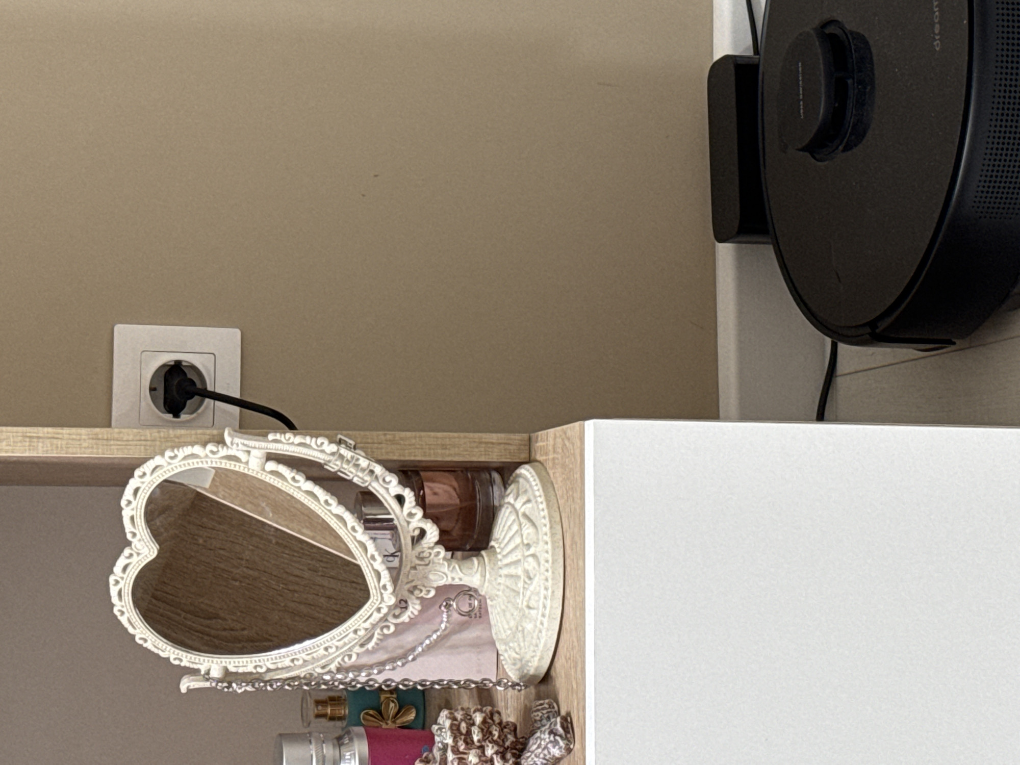 The image shows a section of an interior space, likely a home. On the wall, painted a beige color, is a standard European-style electrical outlet with a black power cord plugged into it. The cord runs along a shelf, which is part of a cabinet. On the shelf, there is a heart-shaped vanity mirror with an ornate white frame. Below the mirror are various items, including perfume bottles and what appear to be pieces of jewelry. To the right of the mirror and cabinet, a black robotic vacuum cleaner is mounted to the wall on a docking station. The vacuum cleaner is partially visible. The location is Unknown, Unknown.