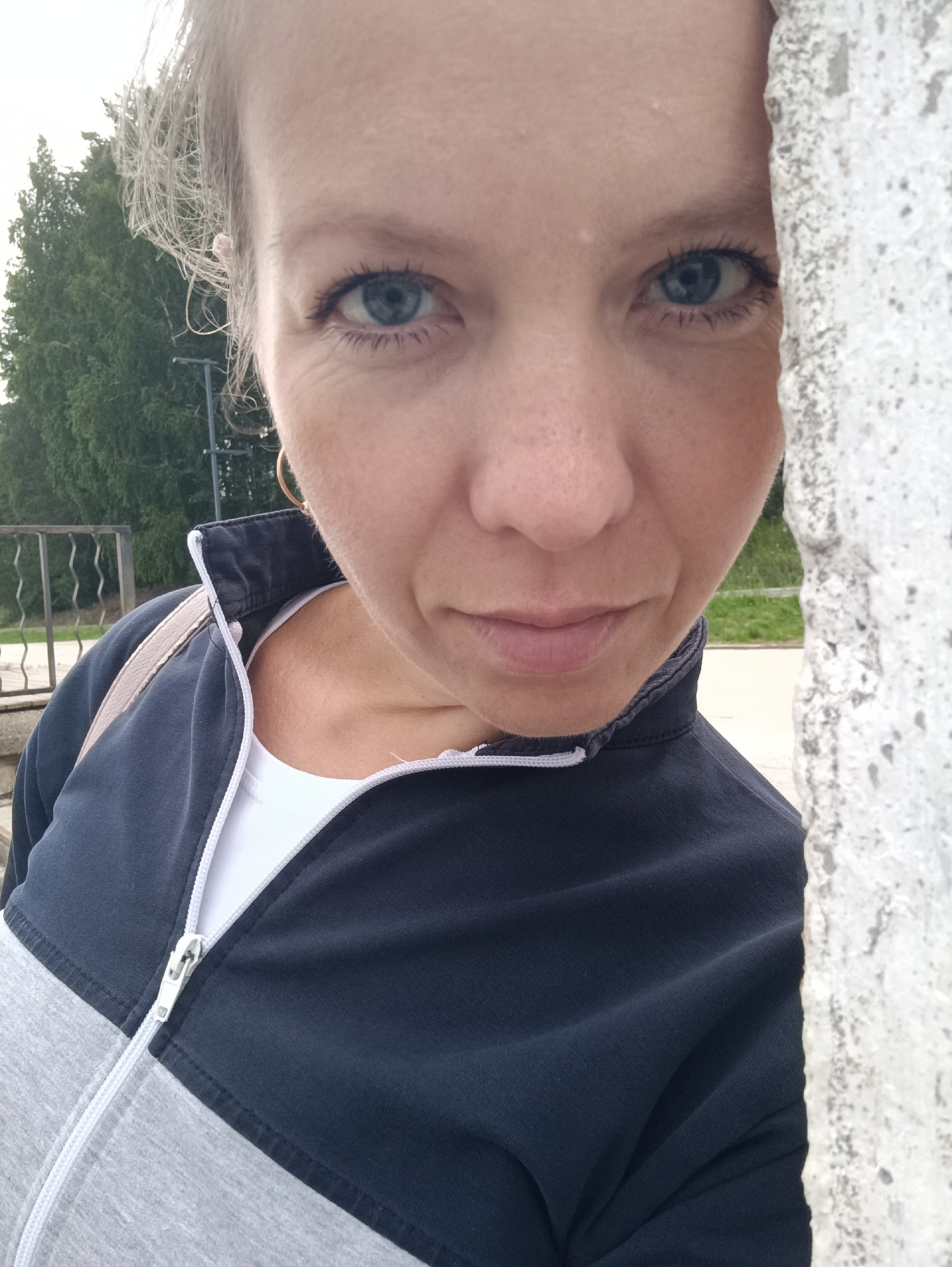 A young woman with light-colored hair and blue eyes is partially obscured by a textured, light-colored wall on the right side of the frame. She is wearing a dark blue and gray zip-up jacket over a white shirt. The background shows green trees and a metal fence, suggesting an outdoor setting, possibly a park or public space within Chaikovsky, Russia. The image is a close-up shot, focusing on the woman's face and upper body.