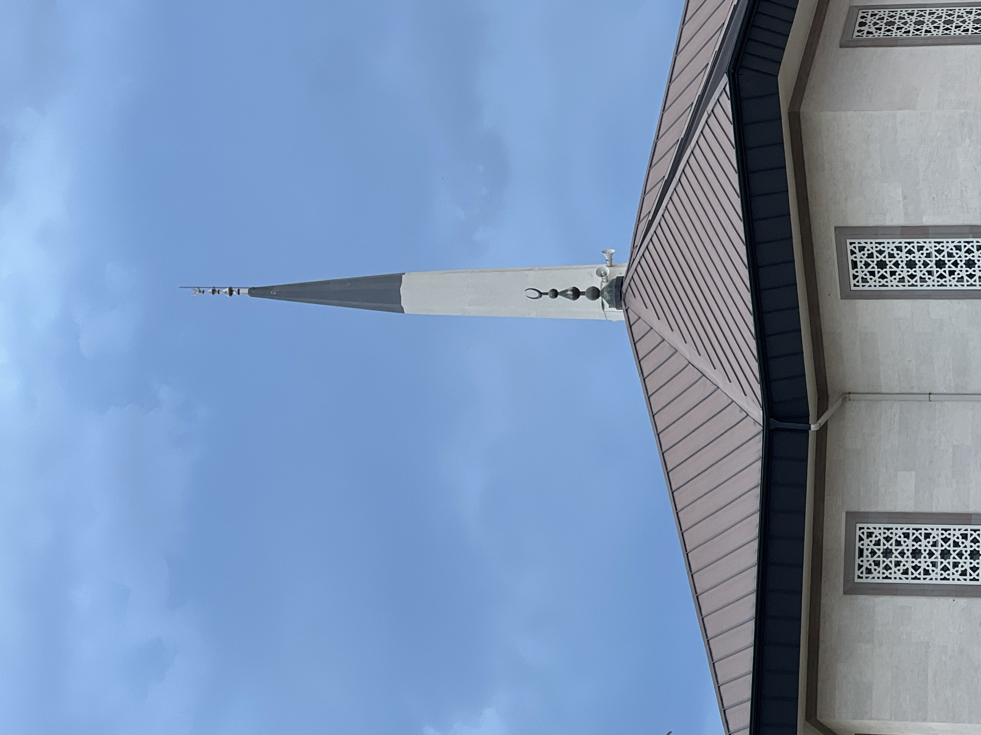 The image shows a mosque's minaret and roof against a cloudy sky. The minaret is tall and slender, tapering to a point, with a dark grey spire at the very top. Below the spire, a crescent moon symbol adorns the minaret, and a speaker is visible attached to its side. The mosque's roof is sloped and appears to be made of metal panels in a brown hue, with a dark, overhang. The building itself is constructed with light-colored stone blocks, and decorative white lattice windows are visible on its facade. The sky is predominantly blue with scattered grey clouds. This mosque is likely the Muratpaşa Mosque, a prominent landmark in Antalya, Turkey, located in the city center. No people are visible in the image.