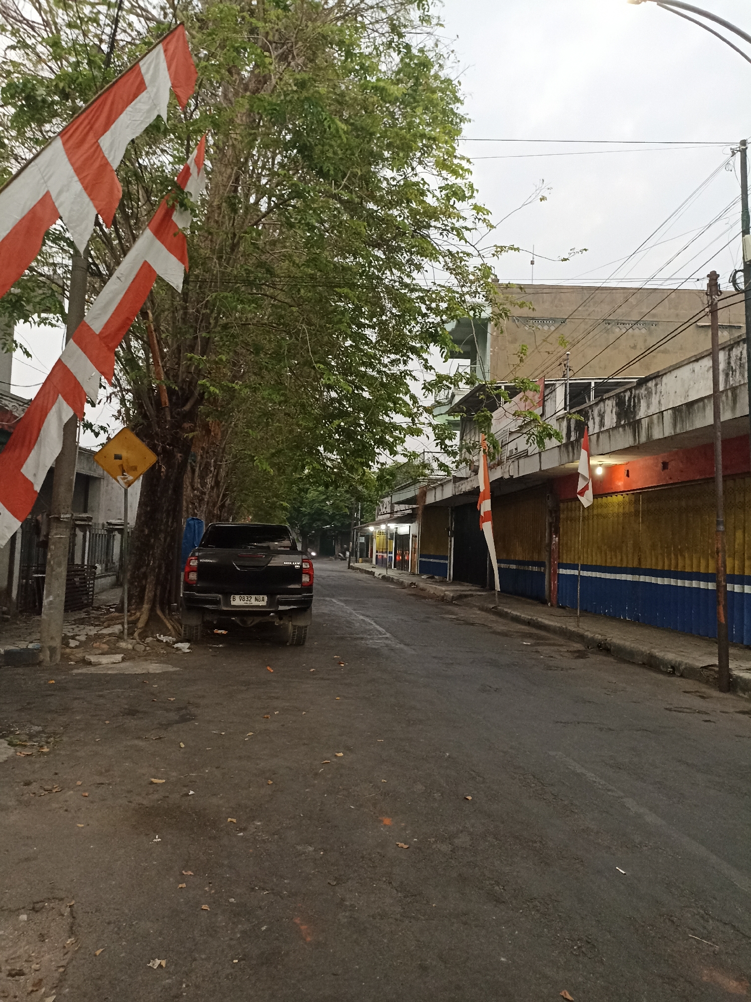 The image depicts a street scene in Lumajang, Indonesia, likely in the morning or late afternoon given the overcast sky. In the foreground, a black pickup truck is parked on the side of a paved road. The truck has a license plate that reads "B 9832 NBA". To the left of the truck, a tall tree with dense green foliage is prominent, its trunk weathered. Several red and white flags, likely Indonesian flags (Sang Saka Merah Putih), are attached to a pole near the tree, their fabric somewhat tattered. A yellow diamond-shaped warning sign is also visible on a pole near the tree.
The road itself is asphalt, with some debris and fallen leaves scattered on its surface. The street curves slightly to the right and recedes into the distance. Along the right side of the road, a row of buildings is visible. These appear to be commercial establishments with shuttered storefronts painted in sections of yellow and blue. Some of these buildings have balconies or upper floors. Two more red and white flags are displayed outside these buildings.
The environment appears somewhat urban but with a strong presence of vegetation. The overall impression is one of a quiet street, possibly before or after business hours. The sky is a muted gray, suggesting diffused light.
