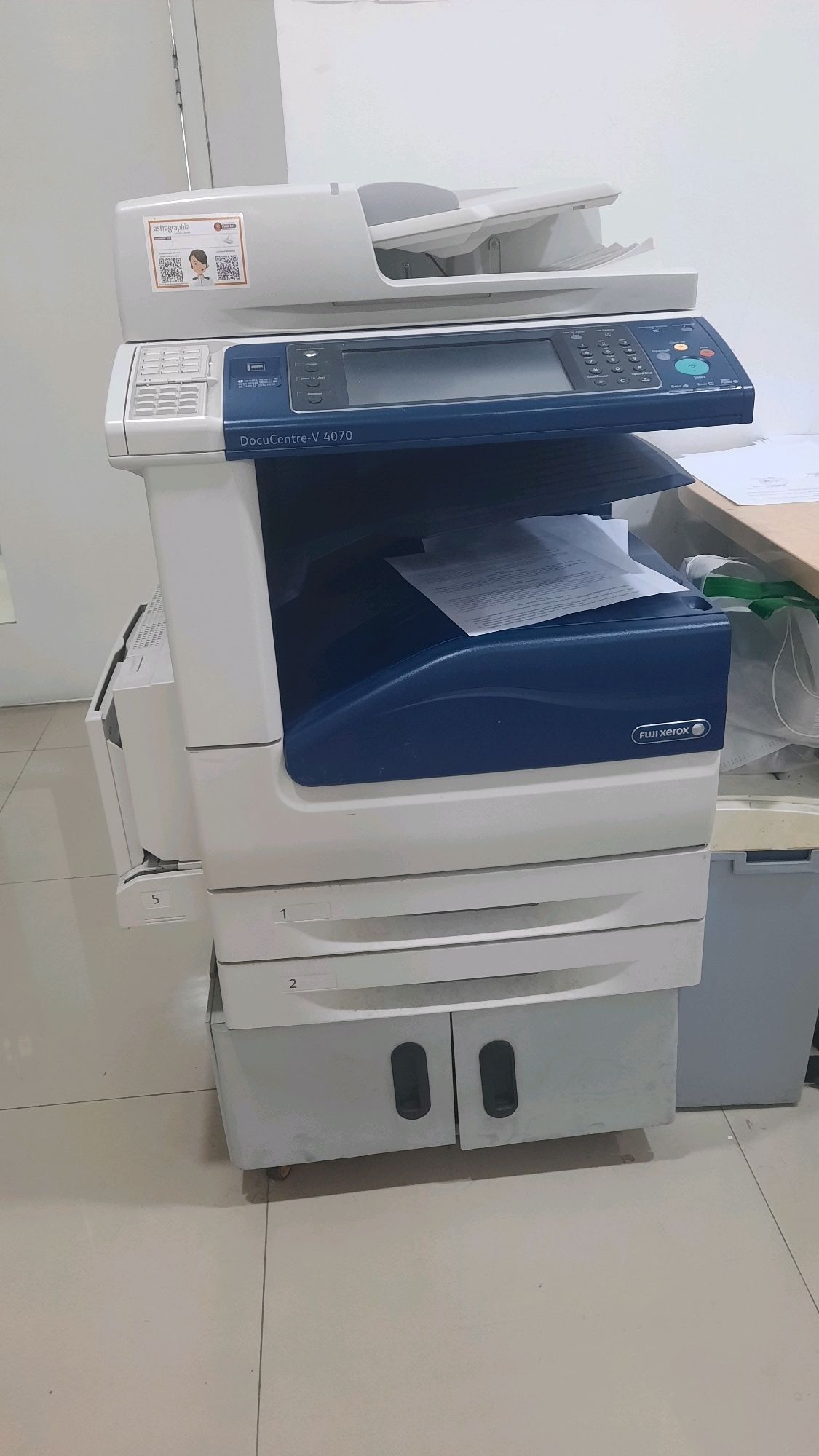 A Fuji Xerox DocuCentre-V 4070 printer is shown in an indoor environment, possibly an office, with tiled flooring. The printer is white and blue, with a scanner at the top and several drawers at the bottom. A document is visible in the scanner's output tray. The printer has a control panel with a screen and buttons. A label on the printer reads "FUJI XEROX". There is also a sticker on the top left that includes the text "astragrahia" and QR codes. The overall scene is well-lit and appears to be a standard office setting.