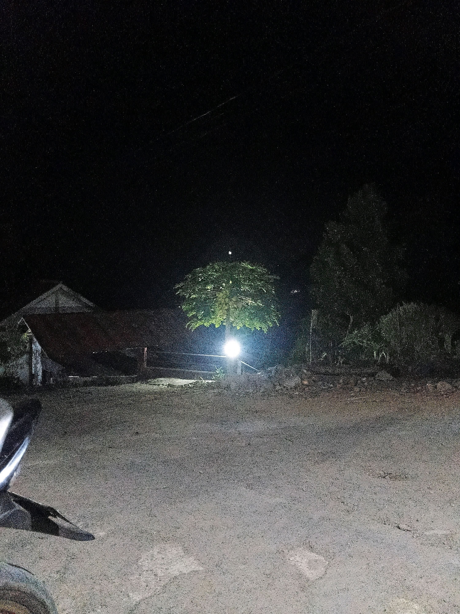 The image depicts a night scene in Sinduaji, Indonesia. A tree is brightly illuminated by a light source, casting a glow on the surrounding area. To the left of the tree, a simple structure with a reddish-brown roof is visible. A dirt road occupies the foreground, with a portion of a motorcycle visible on the left side. The background is dark, with the night sky above, punctuated by a few faint stars. The overall impression is one of a rural or semi-rural setting at night.