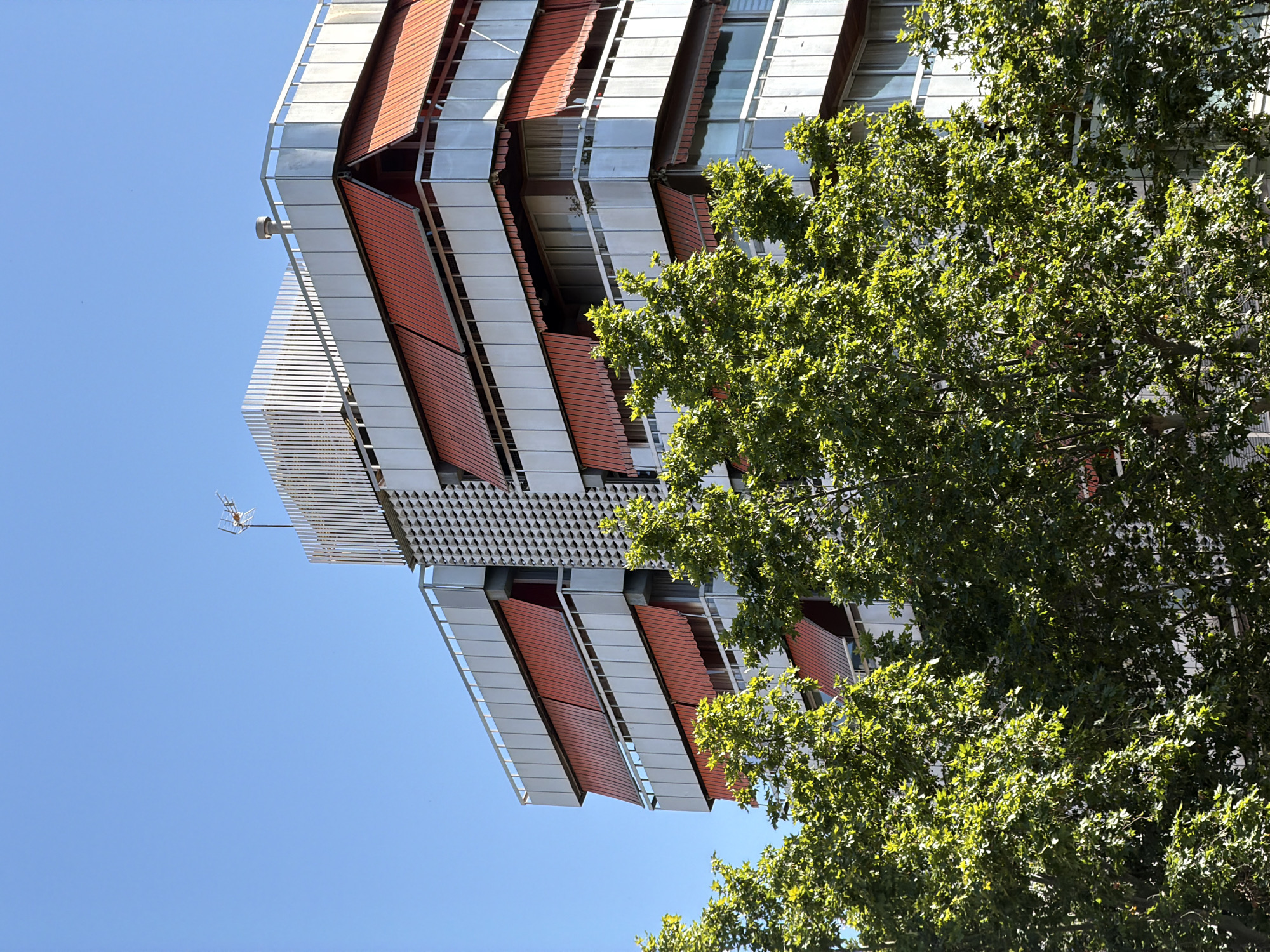 The image shows a low-angle shot of a modern building with a distinctive facade and a large tree. The building is tall and features a repeating pattern of orange and white panels, possibly louvers or shading elements, angled downwards. These are interspersed with sections of glass windows and a metallic lattice-like structure near the top. A small antenna is visible extending from the edge of the building. The sky is a clear, bright blue, suggesting a sunny day. To the right of the building, a lush green tree with dense foliage occupies a significant portion of the frame, its leaves catching the sunlight. The composition is a bit unusual as the image is rotated 90 degrees counter-clockwise, making the building appear to be sideways. This rotation might be a deliberate artistic choice or an error in presentation. The overall impression is one of modern architecture juxtaposed with nature, under clear skies. There is no visible text in the image. Given the context of Barcelona, Spain, this could be a contemporary building in the city. The bright sunlight and clear sky suggest it is daytime, likely mid-morning or afternoon.