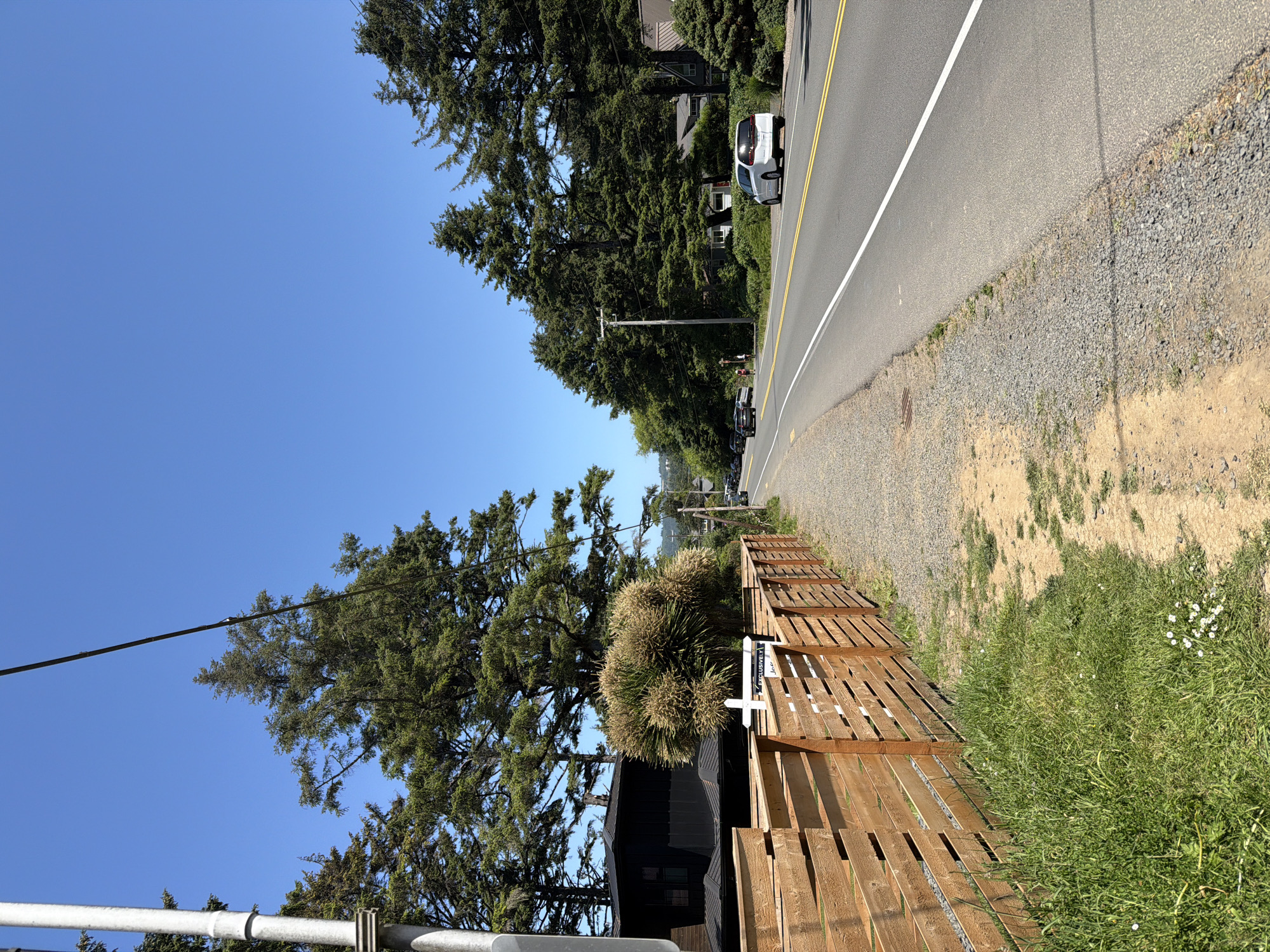 A clear, bright day is depicted in this photograph of a residential street in Cannon Beach, Oregon, United States of America. The road, which runs parallel to a grassy shoulder and a wooden fence, is flanked by tall evergreen trees on both sides. On the left side of the road, a wooden fence encloses a property with a modern, dark-colored building. A white sign with some text is visible behind the fence. On the right side of the road, several cars are parked and driving. A white minivan is in the foreground, and a white sedan is further down the road. The sky is a vibrant blue with no clouds.