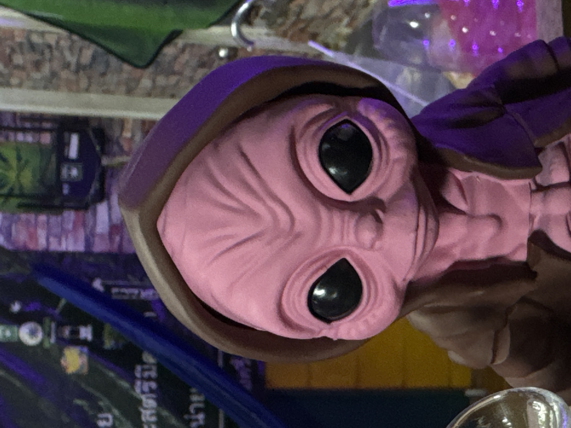 A pink alien figurine is positioned in the center of the frame. The figurine has large, black, almond-shaped eyes, a wrinkled forehead, and a hooded, brown and purple garment. The background consists of a brick wall and a display of items that appear to be posters or decorative pieces, with some glowing purple lights visible on the right side of the image. The lighting is dim, casting shadows on the figurine. The setting is indoors, possibly a room or a display area.