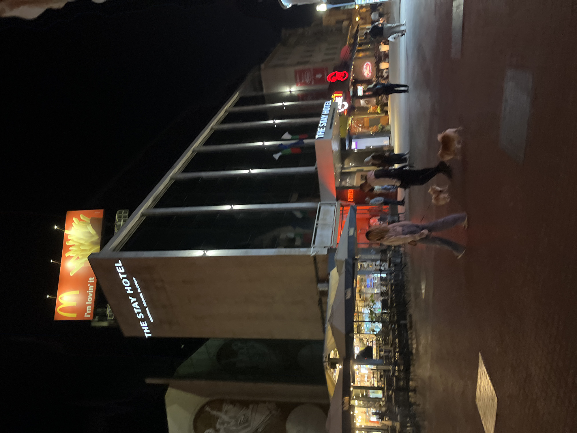 This nighttime image depicts an urban scene, likely in Plovdiv, Bulgaria, based on the presence of "THE STAY HOTEL Central Square Plovdiv" signage. The foreground features the exterior of a building, possibly part of the Stay Hotel, with a large architectural design element. Above, a McDonald's billboard is visible. Several people are walking on the sidewalk in the midground. Tables and chairs are located near buildings in the image. Other signs and lights of adjacent businesses are present. Two dogs appear to be walking on the sidewalk.