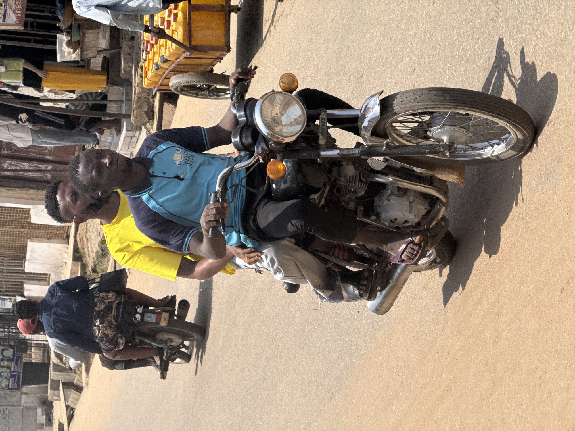 The image depicts a street scene in Abuja, Nigeria, under bright daylight. In the foreground, a man is riding a motorcycle. He is wearing a blue polo shirt with darker blue sleeves and black pants. He is looking forward and appears to be operating the motorcycle with both hands on the handlebars. Behind him, another person is visible on the motorcycle, wearing a yellow shirt and dark pants. Their heads are turned slightly away from the camera.
In the middle ground, another motorcycle is seen from the rear, carrying two passengers. The rider, wearing a red cap and a dark denim jacket, is seated in front. Behind the rider, a person wearing a patterned dress is seated, holding onto the rider. They both have bags.
Further in the background, on the right side of the image, a yellow cart is being pushed by an unseen person. The cart is filled with large, yellow plastic containers, possibly for carrying liquids. In the far background, buildings and street infrastructure are visible, including what appears to be a market stall with clothing on display. The road surface is paved and appears dusty. The overall scene suggests a busy urban environment with people engaged in daily activities like transportation and commerce.