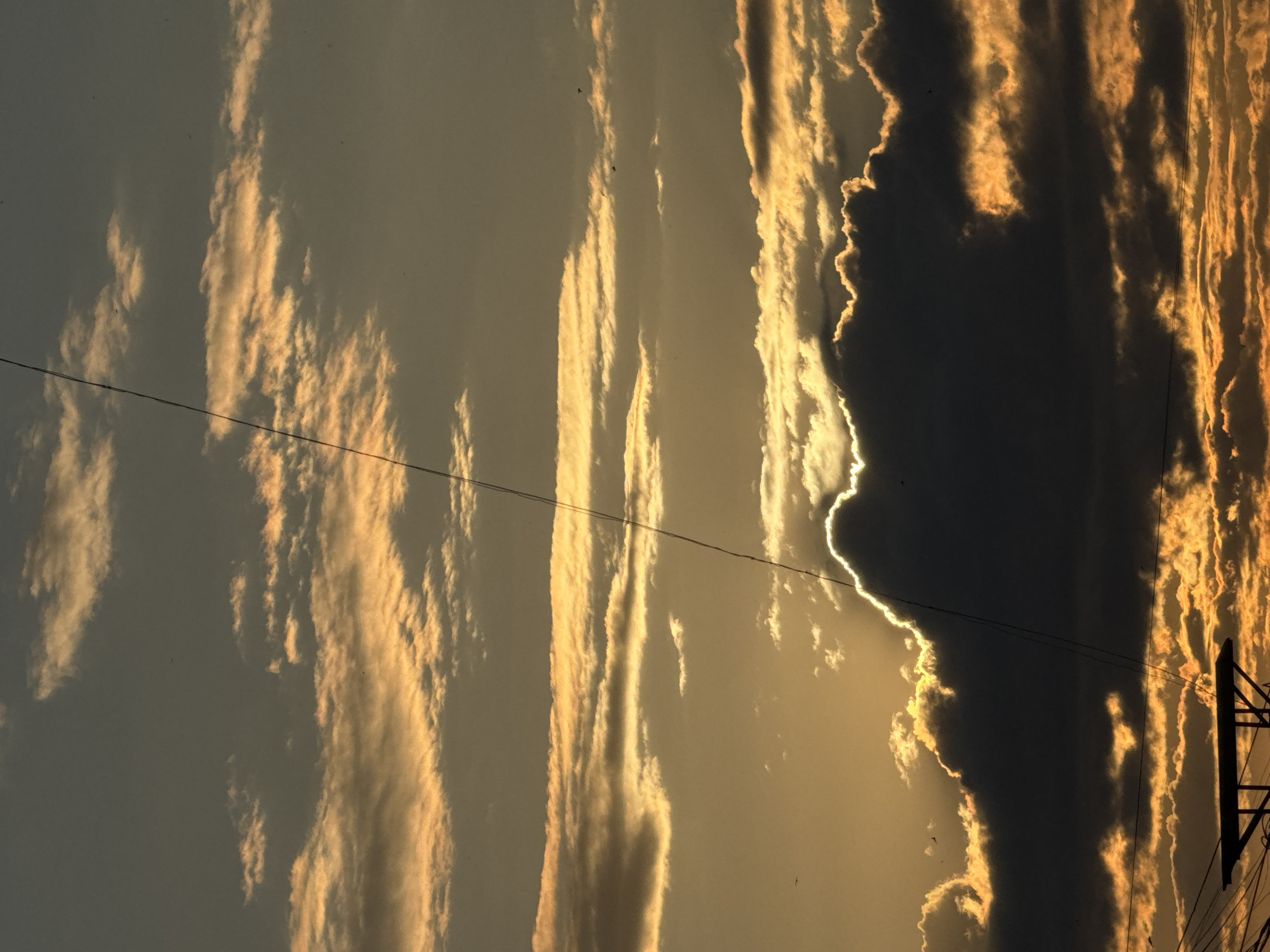 The image depicts a sunset over the city of Chaikovsky, Russia. The sky is filled with dramatic clouds illuminated by the setting sun. The clouds are a mix of dark gray and bright orange, with the sun peeking through some of the darker clouds, creating a halo effect. A thin, dark power line runs diagonally from the top left to the bottom right of the frame, bisecting the sky and clouds. In the bottom portion of the image, a silhouette of a power line structure with horizontal and diagonal supports is visible against the bright, cloud-filled sky. The scene is tranquil and captures the natural beauty of the sky at dusk.