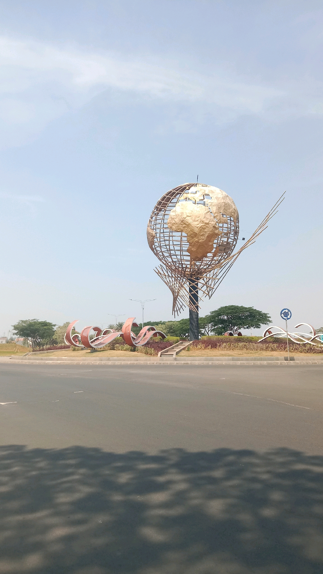 A globe sculpture stands prominently in a roundabout in Telagamurni, Indonesia. The sculpture features a large, golden globe with a lattice-like structure supporting it, appearing to represent a stylized representation of Earth. In front of the globe, an abstract red sculpture with curved, wavy elements is visible, adding an artistic touch to the landscape. The roundabout itself is surrounded by green grass and some trees, with a clear sky overhead. The foreground shows a paved road with the shadow of trees cast across it, indicating a sunny day. A blue sign with a white arrow indicating a roundabout is also visible to the right of the sculpture. The overall scene depicts a public space with decorative elements designed to be aesthetically pleasing and perhaps serve as a landmark.