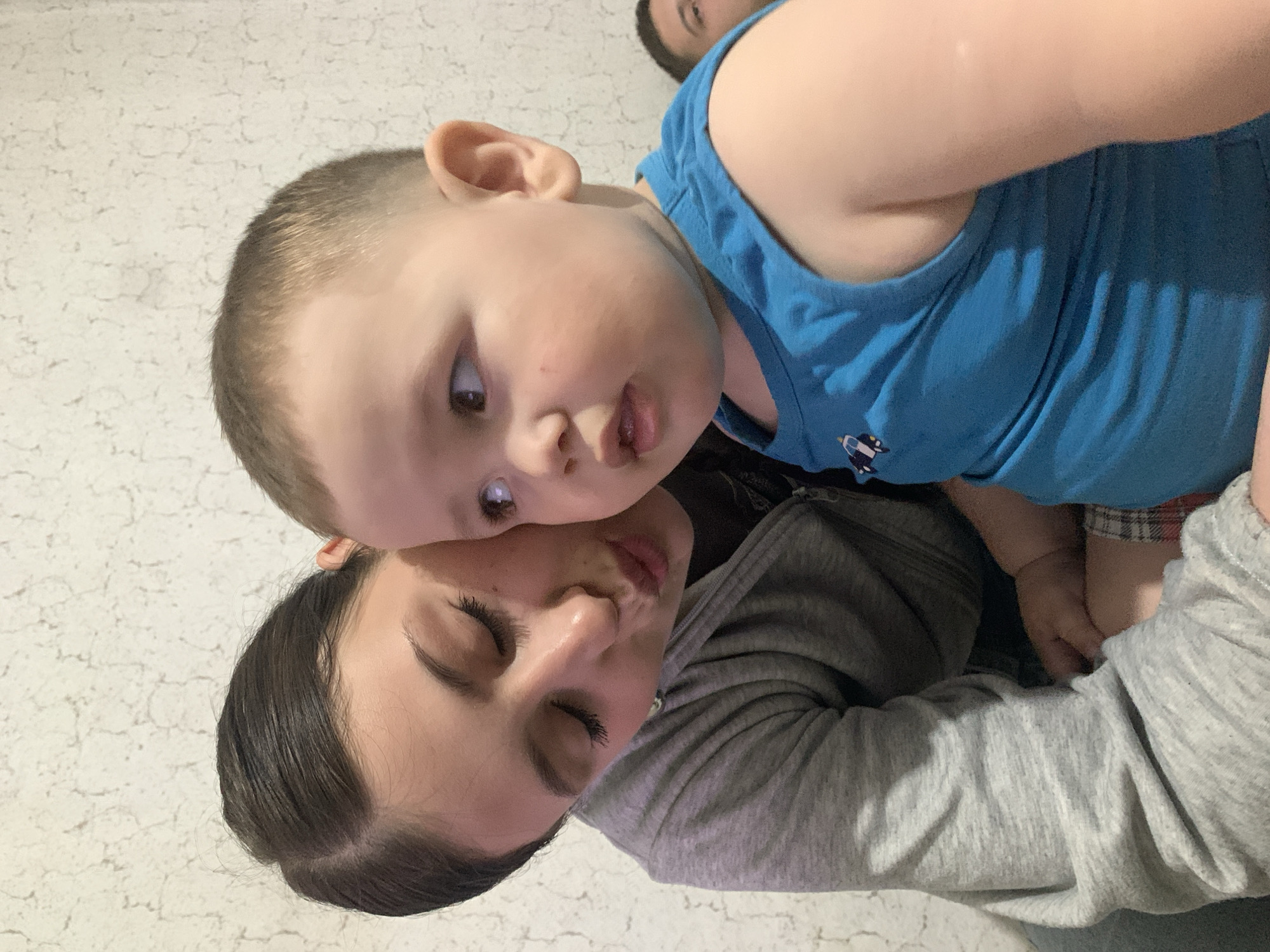 A young woman in a gray sweatshirt is hugging a young boy in a blue tank top. The boy's head is near her cheek, and his eyes are open. The background is a white textured wall. Another person's head is visible at the very top of the image. The location is unknown and there is no evidence in the image to suggest this took place in Фоки, Russia.