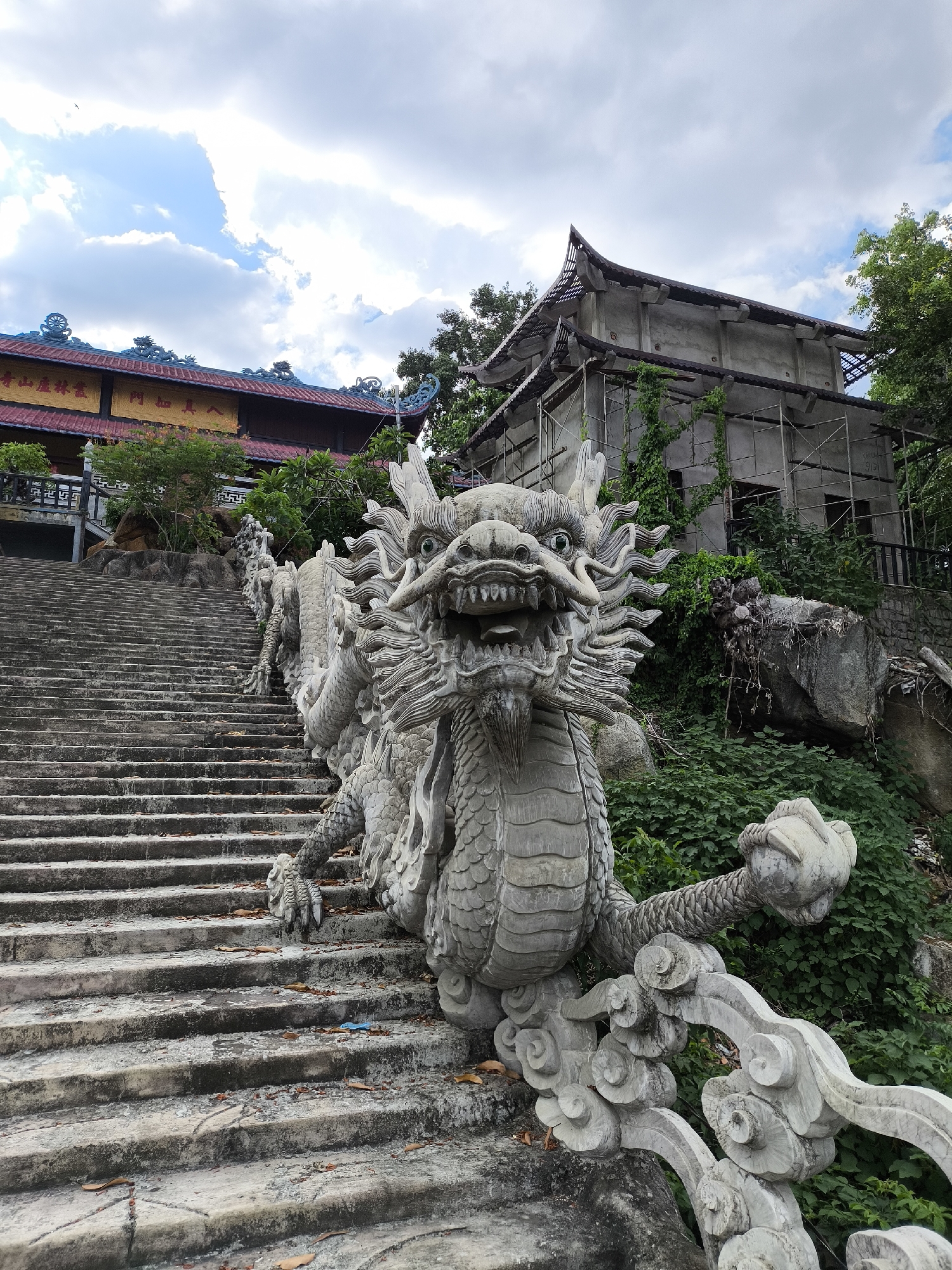 The image depicts an outdoor scene at a temple in Nha Trang, Vietnam. The foreground is dominated by a stone dragon statue, intricately carved, positioned alongside a long, wide stone staircase. The dragon is depicted in a coiled posture, its head facing forward with an open mouth and sharp teeth, its body extending along the side of the steps. The staircase itself is weathered and shows signs of age, with leaves scattered on some of the steps.
Behind the staircase, to the left, is a building with a red roof, featuring traditional East Asian architectural elements. It has a blue-tiled roof edge and ornate decorations. Yellow signage with Asian characters is visible on the building's facade. To the right of the staircase, a partially constructed building is visible, covered in scaffolding and overgrown with green vines. This suggests that the temple complex may be undergoing renovation or expansion.
The setting is lush and green, with dense foliage surrounding the buildings and the stairs. Large rocks are also present on the right side of the image, integrated into the natural landscape. The sky above is partly cloudy, with a mix of blue sky and white clouds, indicating it is daytime. The overall impression is one of a serene, spiritual location, with elements of both ancient tradition and ongoing development. No people are visible in the image.