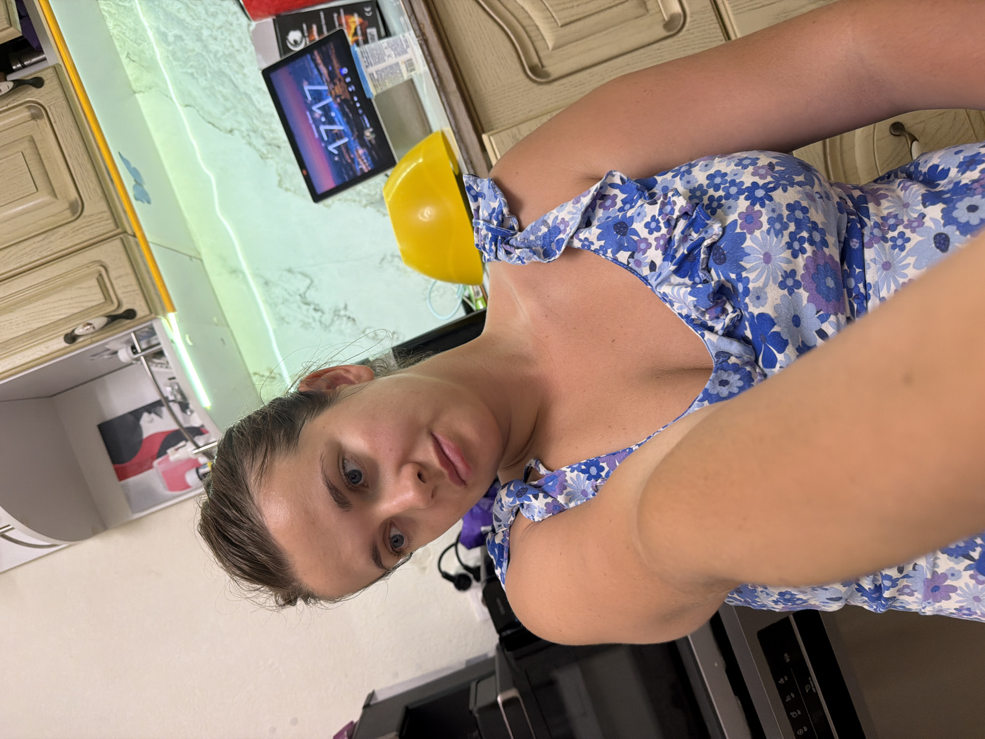 In Sochi, Russia, a young woman with brown hair pulled back and blue eyes is pictured from the chest up, looking to her left. She is wearing a blue and white floral print dress with ruffled straps. Her right arm is extended towards the camera, slightly blurring the foreground. The background suggests a kitchen environment with cream-colored cabinets, a yellow object, and a tablet displaying the time 17:17.