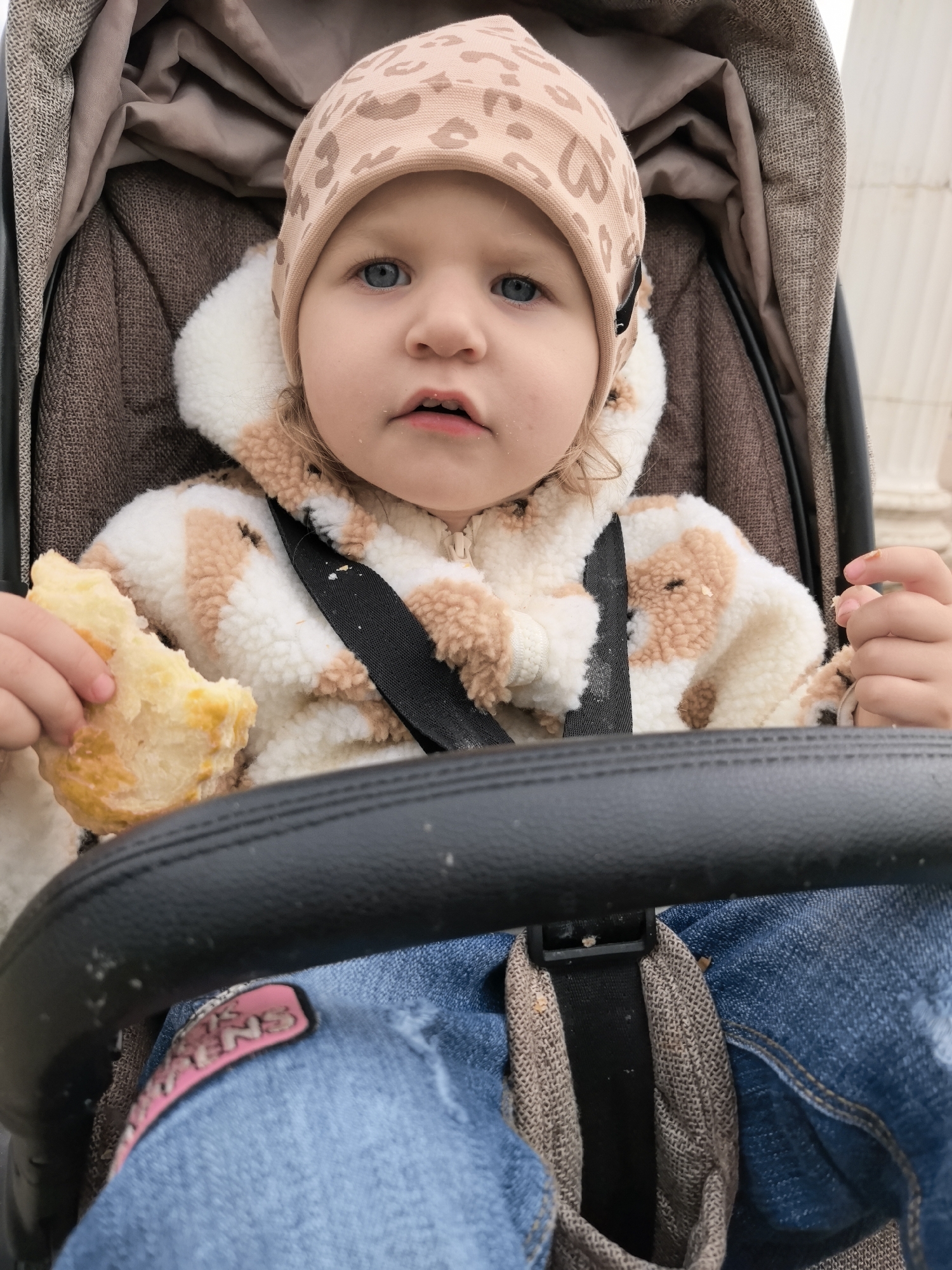 A toddler with blue eyes is sitting in a stroller. They are wearing a beige hat with a leopard print, a white fleece jacket with beige spots and a hood, and blue jeans with a pink patch on the left leg. The toddler is holding a piece of yellow bread in their left hand. The stroller has a brown fabric interior, a black safety bar, and a black seatbelt. The location could be the city of Chaikovsky, Russia.