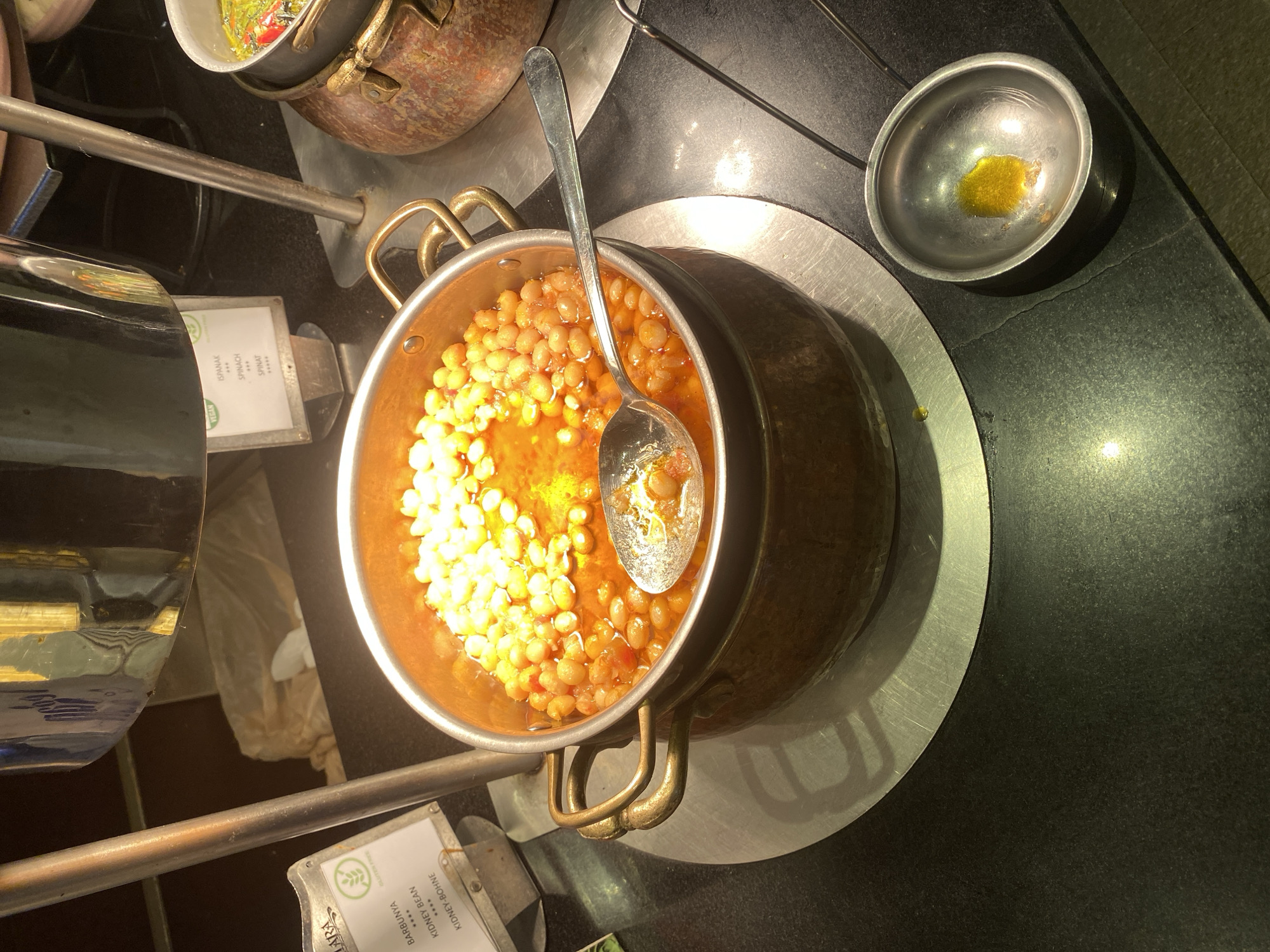 This image shows a close-up of a buffet in Aksu, Turkey. The primary subject is a copper pot filled with a stew of chickpeas and beans in a red sauce. A silver spoon rests on the edge of the pot, with some stew on it. To the left of the pot is a small sign with the words "BARBUNYA," "KIDNEY BEAN," and "KIDNEY-BOHNE." To the right of the main pot, there is another smaller copper pot containing what appears to be pickled vegetables and chili peppers. In the foreground, a small silver dish with a yellowish liquid, possibly oil, is visible. The background is dark and out of focus, with reflections of light on metallic surfaces. The scene suggests a restaurant or food service environment where these dishes are being served.
