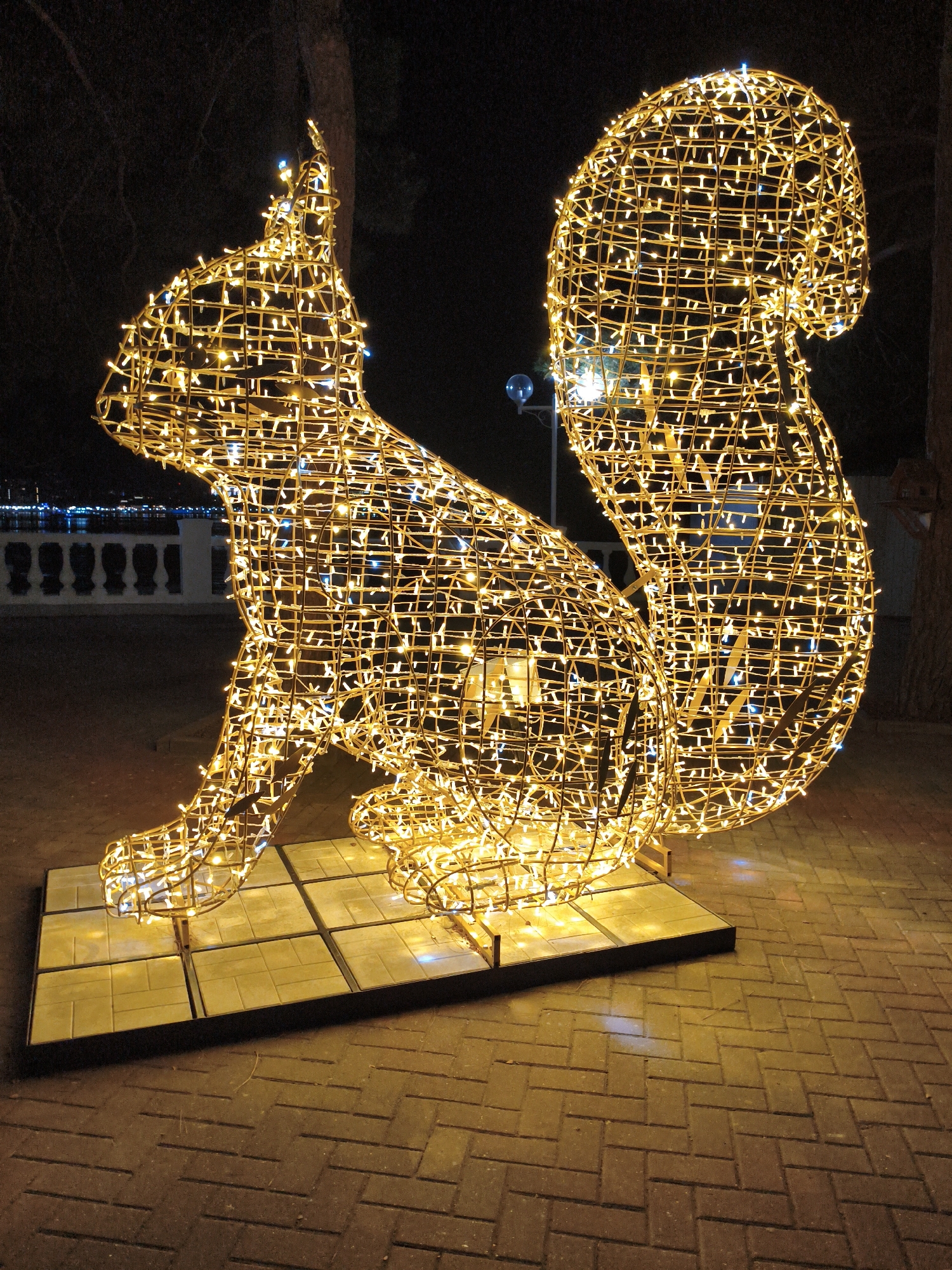 The image depicts a large, illuminated squirrel sculpture made of wire and adorned with numerous yellow LED lights. The sculpture is situated outdoors at night on a paved walkway. The squirrel is posed in a seated position, with its bushy tail curled up behind it. It rests on a rectangular platform constructed of light-colored square tiles.
In the background, there are faint indications of a waterfront area with lights reflecting on the water. A white balustrade or railing is visible along the water's edge, and beyond that, distant lights suggest a town or city. A lamppost with a blue light is visible to the left of the squirrel. A tree trunk is partially visible on the right side of the frame. The overall scene suggests a festive or decorative display, possibly for a holiday season. The time of day is clearly night, as indicated by the dark sky and the illumination provided by the sculpture and background lights. The weather appears clear, with no visible rain or fog. There is no visible text in the image. The setting is likely a public space, such as a park or promenade.