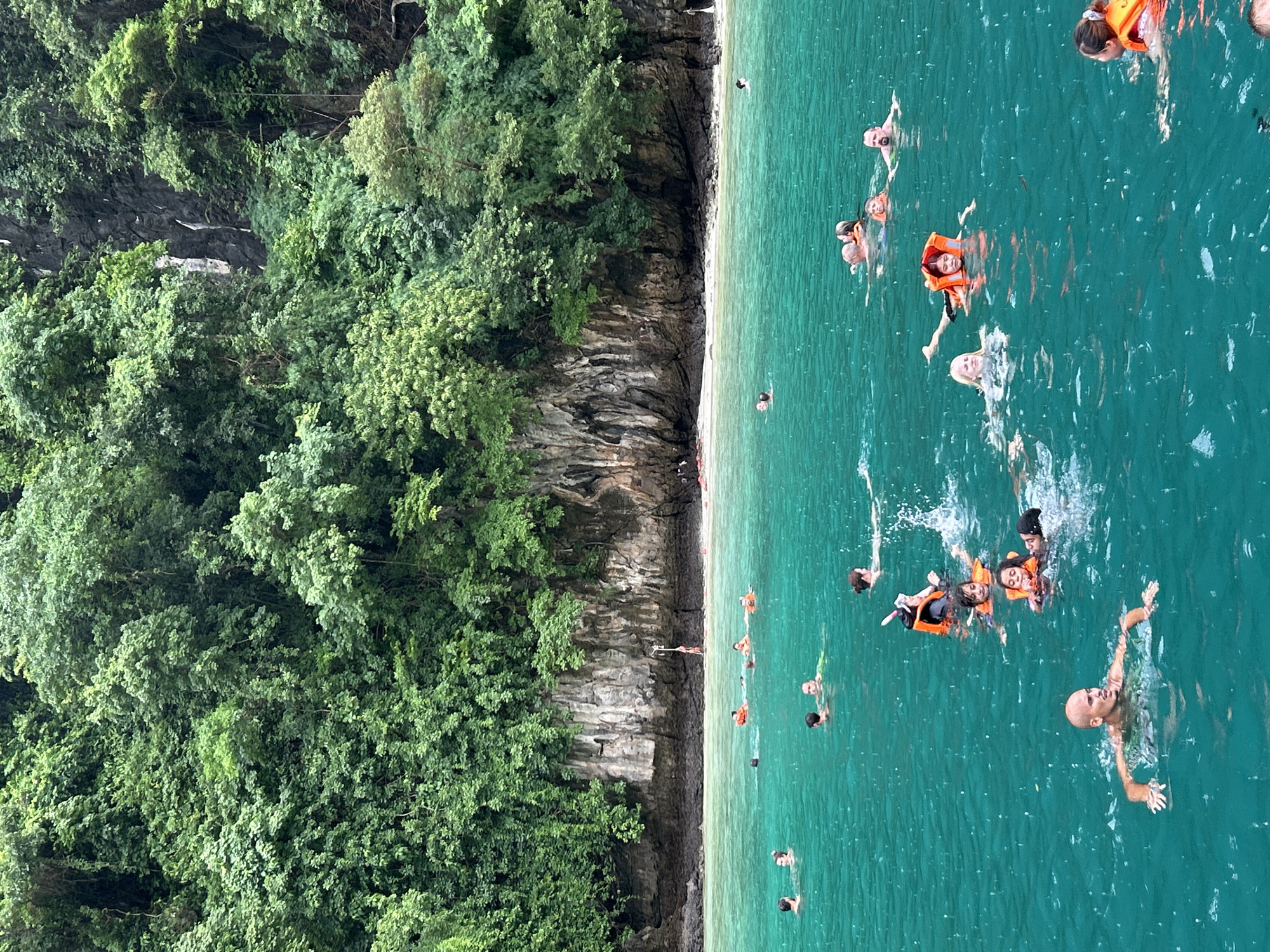 This image captures a vibrant scene in the waters off Ao Nang, Thailand, showcasing a group of people enjoying a swim near a lush, green, forested cliff face. The water is a striking turquoise, with visible ripples and droplets indicating either a recent rain shower or an active group of swimmers.
In the foreground, several individuals are visible. A bald man with tanned skin is swimming towards the viewer, his arms outstretched and his gaze directed forward. To his right, a woman with blonde hair and an orange life vest is swimming, her expression neutral. Further back, a person wearing an orange life vest and a snorkel mask is splashing water, appearing energetic. Several other individuals are scattered throughout the water, some wearing orange life vests, suggesting safety precautions.
The background is dominated by a steep, rocky cliff densely covered in vibrant green vegetation. The foliage appears thick and untamed, characteristic of tropical landscapes. The cliff face shows layers of rock and soil, with some areas appearing lighter due to the lighting. A small sandy beach is visible at the base of the cliff, separating the water from the dense greenery. In the distance, a few more figures can be seen swimming or wading near the shore. The overall atmosphere is one of recreation and natural beauty, with the activities of the people harmoniously blending with the impressive natural environment.