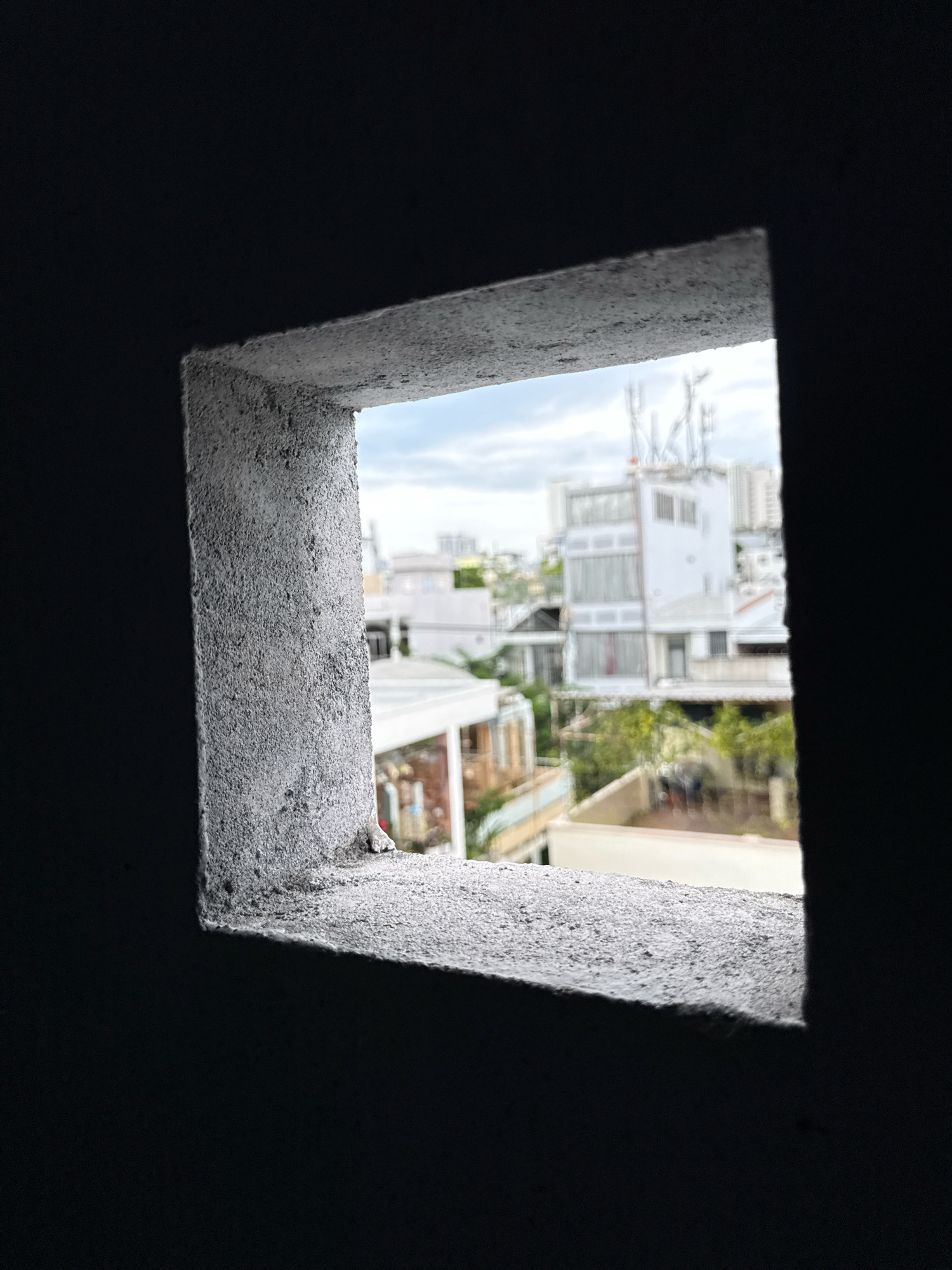 This is a photograph taken from inside a dark, enclosed space, looking out through a small, rough, concrete square opening. The opening frames a view of an urban environment in Nha Trang, Vietnam. The foreground is dominated by the textured, gray concrete of the window frame, which appears weathered and unfinished. The interior of the space is dark, creating a strong contrast with the brighter scene outside.
The view through the opening reveals a dense collection of buildings, characteristic of a Vietnamese city. White and light-colored buildings are prominent, some appearing to be multi-story residential or commercial structures. One building in the middle ground has several antennas or communication towers on its roof. Lush green foliage, likely trees and plants, can be seen interspersed between the buildings, suggesting a tropical climate. The sky is overcast with clouds. The overall impression is one of looking out from a sheltered or hidden vantage point onto a bustling city street or neighborhood. There are no clearly discernible people or specific activities visible in the frame.