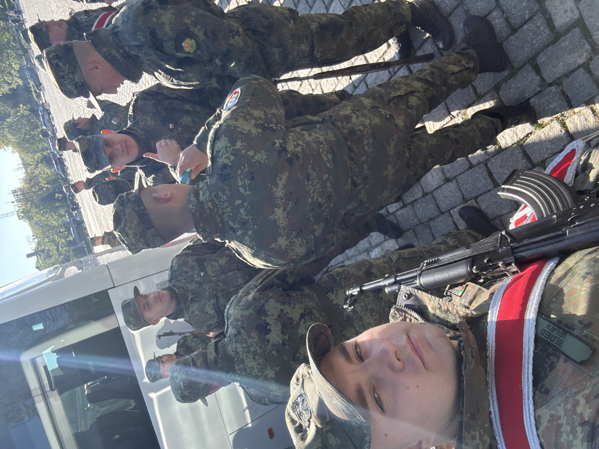The image shows a line of soldiers wearing camouflage uniforms and caps, likely in Sofia, Bulgaria. They are standing on a paved surface, possibly a square or street, with a white vehicle partially visible to their left. The soldiers appear to be preparing for an event or parade, and one of them is holding an AK-pattern assault rifle. A soldier in the front is looking at the camera, displaying his name badge. The background shows some buildings and trees, indicating an urban setting.