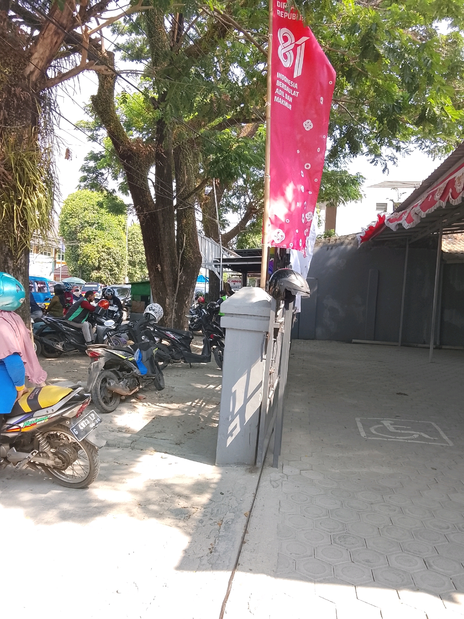 A street scene in Slawi, Indonesia, captures a collection of parked motorcycles and a few individuals under a canopy of trees. A prominent red banner with white lettering, possibly commemorating an Indonesian national holiday, hangs on a pole. In the foreground, several motorcycles are visible, one with a yellow seat and a license plate reading "G 4973 LP". A woman wearing a pink headscarf and a blue garment stands near the front of a motorcycle. To the right, a concrete pathway with hexagonal tiles leads towards a building with a red and white striped awning. A symbol for disabled access is marked on the pavement. The overall atmosphere suggests a public space, likely a market or street side area, with vehicles and pedestrian activity.