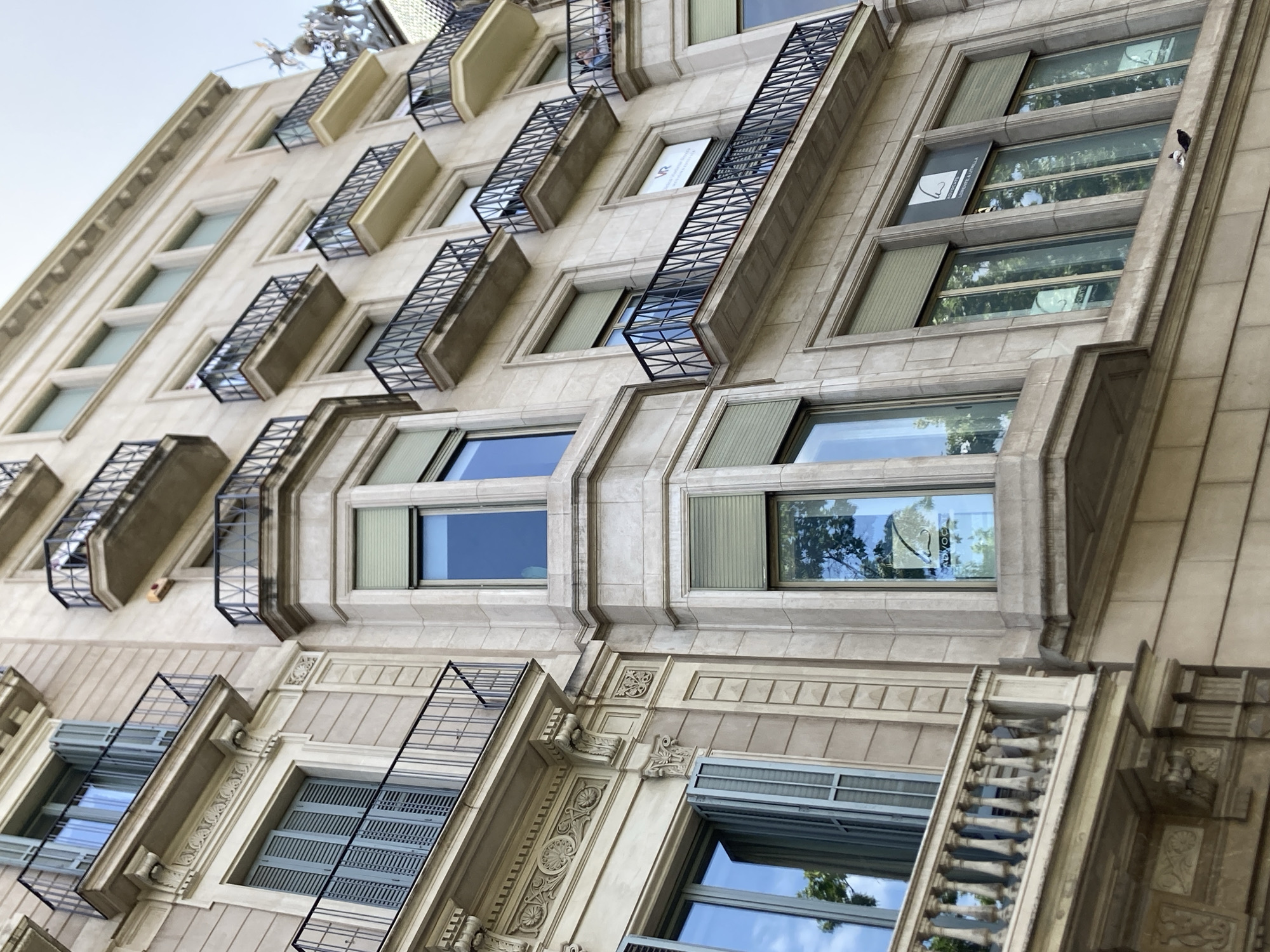 This is a low-angle, close-up, outdoor shot of an ornate, historic building in Barcelona, Spain. The building appears to be made of light-colored stone and features multiple stories with numerous windows. The windows are of various sizes and styles, some with decorative metalwork or grates, and others with blinds. The architecture suggests a turn-of-the-century or early 20th-century style, with detailed moldings and stonework. Several windows have signs displayed on them, one of which shows a logo that appears to be in a stylized "S" or "R" shape with additional abstract elements. Another sign is partially visible with text that might read "LOIXEL I LA CINTRA". The reflections in the windows show trees and sky, indicating it is daytime and likely clear weather. The overall impression is of a well-preserved, elegant building in an urban setting. There are no people visible in the image, but the presence of balconies and windows suggests residential or commercial occupancy.