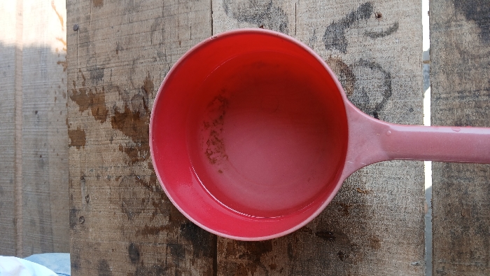 A red plastic ladle, partially filled with a clear liquid containing dark particulate sediment, rests on a weathered wooden surface. The wooden surface comprises multiple planks, displaying visible grain, discoloration, and scattered water droplet stains. In the upper left quadrant, a portion of a light-colored fabric, consistent with a white or light blue garment, is marginally visible. Bright natural light illuminates the scene from the upper right. The observed location is in the city of Bandasafa, Indonesia. No direct human presence or interaction with the ladle is fully discernible.