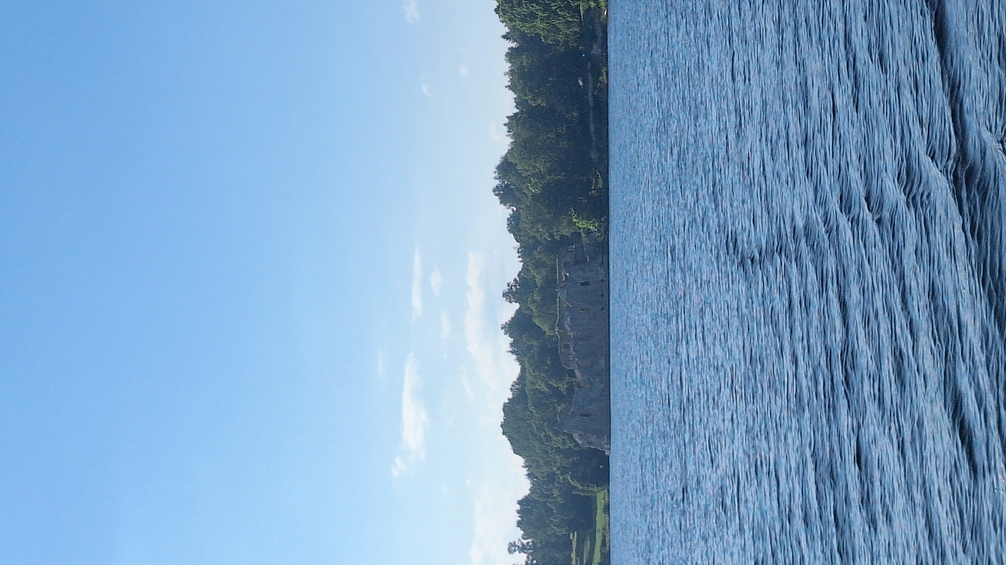 The image depicts a serene waterscape with a ruined structure on the shore. The foreground is dominated by the rippling surface of a body of water, likely a lake or a wide river, with gentle waves creating patterns of light and shadow. The color of the water is a deep blue, reflecting the clear sky above.
In the middle ground, a dense line of green trees forms a backdrop against the sky, suggesting a wooded area. Nestled within this foliage, and extending towards the water, are the remains of a stone structure. The ruins appear weathered and ancient, with irregular shapes and openings visible, hinting at a historical building, possibly a castle or fortification. The stone is a muted gray, contrasting with the vibrant green of the surrounding trees.
The sky above is a brilliant, cloudless blue, indicative of a sunny day. The overall atmosphere is one of tranquility and natural beauty, with a touch of historical mystery provided by the ruins. The scene is devoid of any human presence or activity, emphasizing the quietude of the location.
This scene is likely located in the vicinity of Sece, Latvia, known for its natural landscapes and historical sites, which might include such ruins.