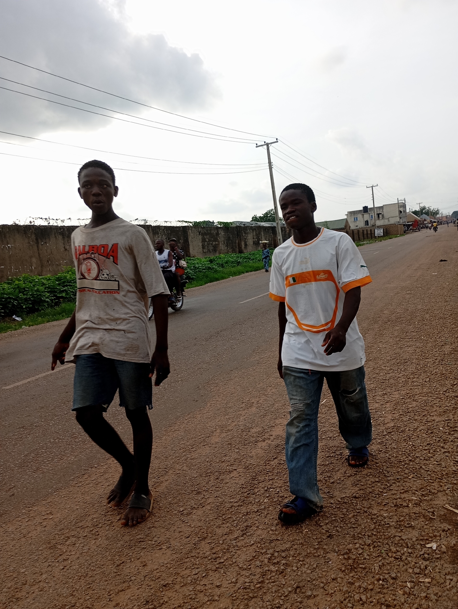 Two young males are walking on the right side of a paved road in Shendam, Nigeria. The male on the left is wearing a light-colored, stained t-shirt with graphics and dark denim shorts. He is barefoot on his right foot and wearing a sandal on his left. The male on the right is wearing a white t-shirt with orange trim and blue ripped jeans, along with blue sandals. In the background, there is a concrete wall with barbed wire on top, followed by dense green foliage. Further down the road, there are buildings, utility poles, and a few other individuals, including two people on a motorcycle. The sky is overcast with scattered clouds.