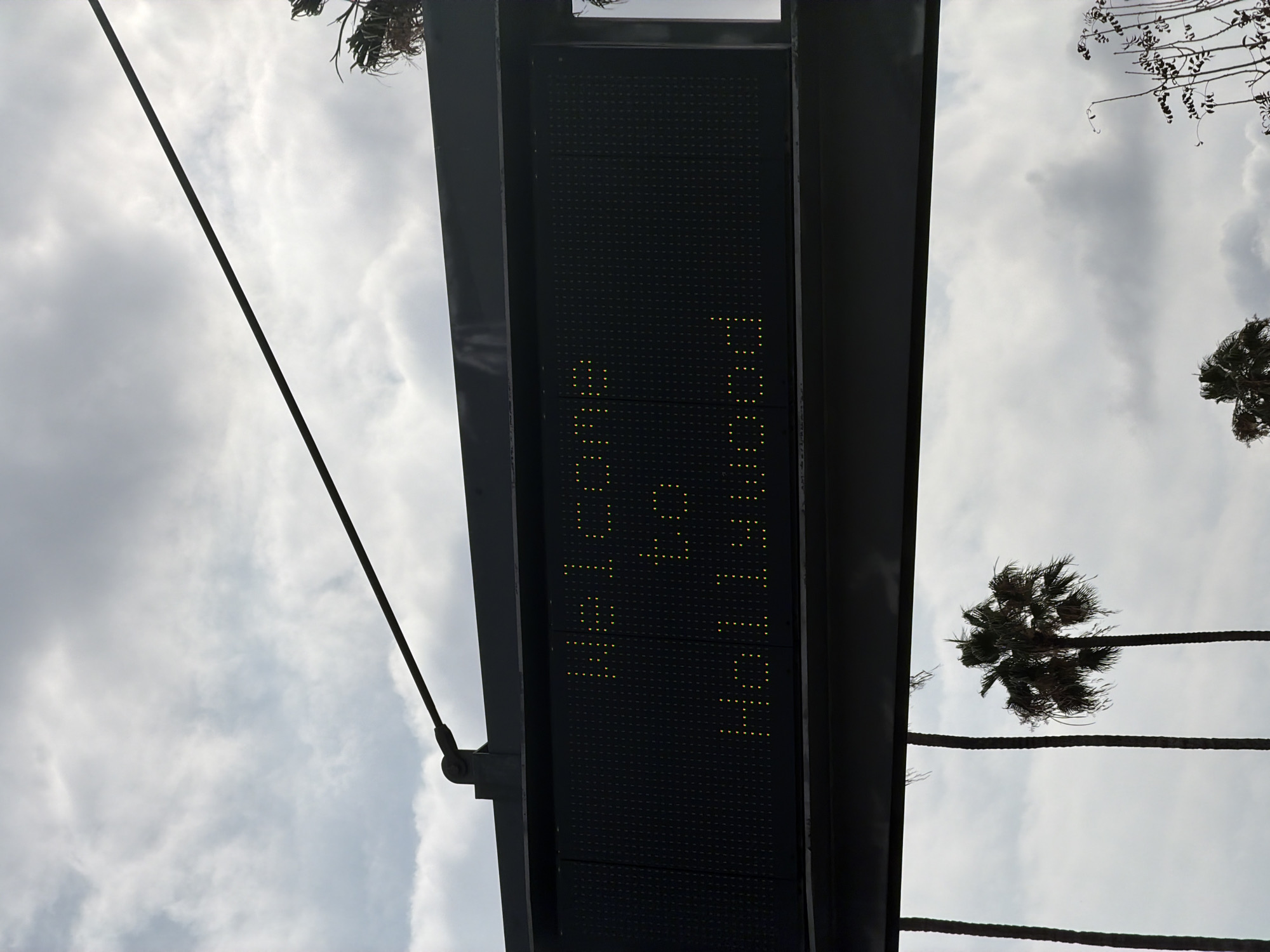 The image shows an electronic sign against a cloudy sky in what appears to be Los Angeles, USA, based on the presence of palm trees. The sign displays the text "Hollywood 6:29 AM". The sign is framed by a dark metal structure. A support cable is visible on the left side of the frame.