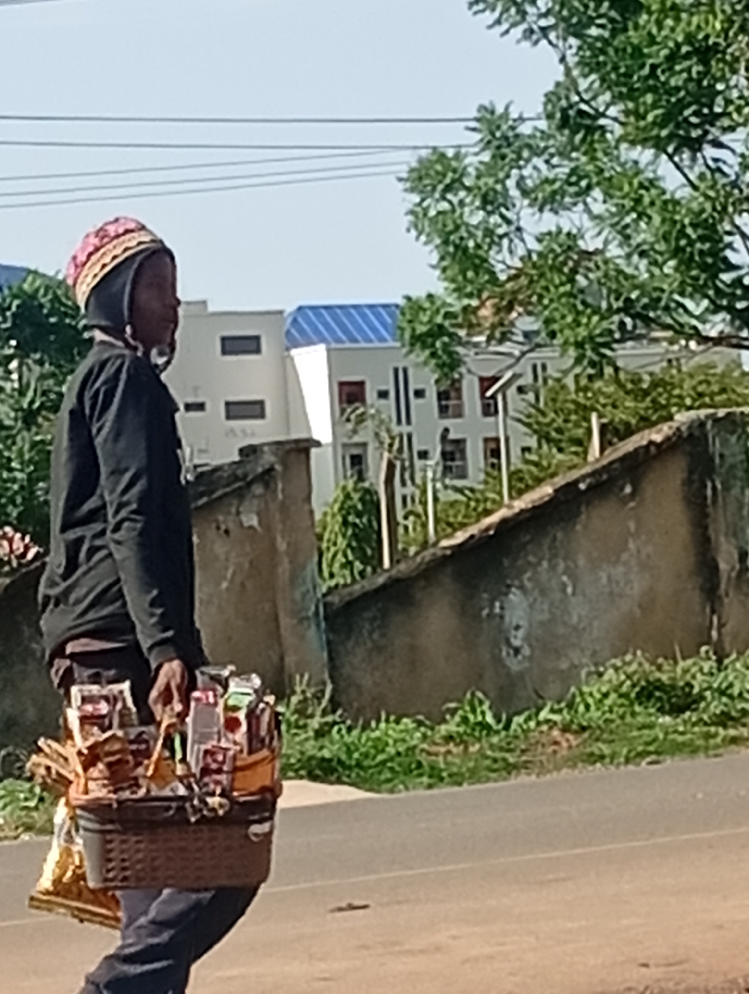 A person walks along a street in Rafin Par, Nigeria, carrying a woven basket filled with goods, likely for sale. The person wears a black hooded sweatshirt and a patterned hat. The basket contains numerous small packaged items, possibly snacks or toiletries, and some larger, golden-wrapped items. The background shows a textured wall, lush green vegetation, and modern buildings with white facades and blue roofs. The sky is clear and pale. The street appears to be paved with asphalt.