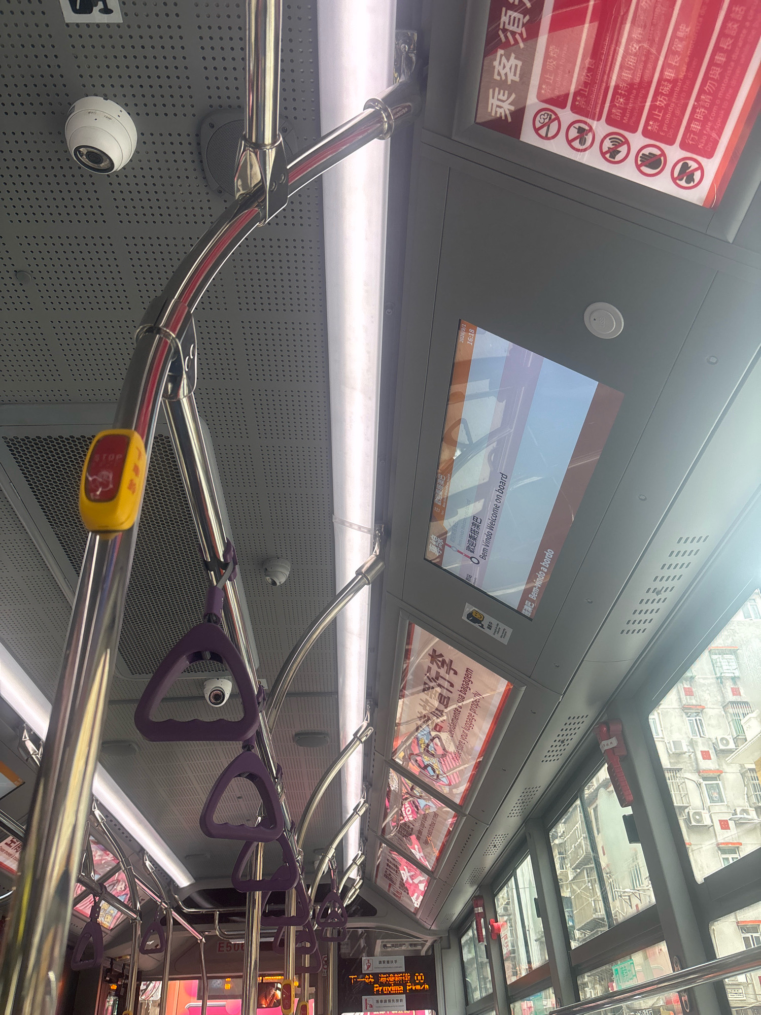 The image shows the interior of a public bus in Macau, China. The perspective is from a passenger looking upwards and towards the front of the bus. Prominently visible are the chrome handrails and purple grab handles for standing passengers. A white security camera is mounted on the ceiling. To the right, a digital screen displays a welcome message in multiple languages, including "Bem-vindo Welcome on board" and "Bem-vindo a bordo". Above the screen, a red and white sign features several prohibition symbols, such as no smoking, no eating, and no littering, in Chinese and possibly Portuguese. The ceiling is a textured grey material with numerous small perforations. On the left side, a yellow stop request button with a red flashing light is attached to a handrail. The windows on the right side of the bus offer a glimpse of buildings and air conditioning units typical of urban Macau. The lighting suggests it is daytime, with sunlight entering through the windows.