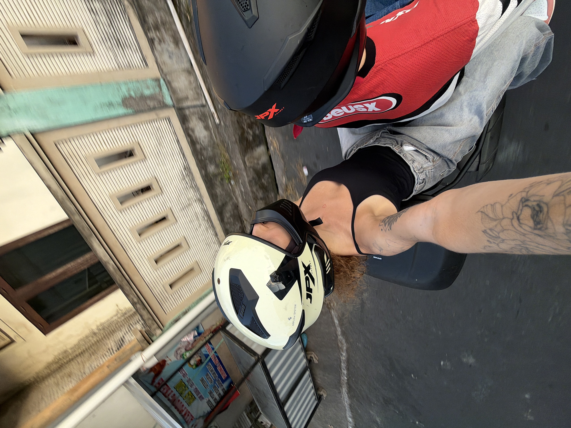 Two individuals are riding a motorcycle down a street in North Kuta, Indonesia. The individual in the foreground is wearing a white and black JPX helmet, a black tank top, and light-wash denim jeans. They have tattoos on their right arm. The person behind them is wearing a black helmet and a red and white jersey. The environment appears to be urban, with buildings lining the street. One building has a sign with Indonesian writing. The road is paved and appears wet in places. The image is taken from a first-person perspective, as if from the rider of the motorcycle.