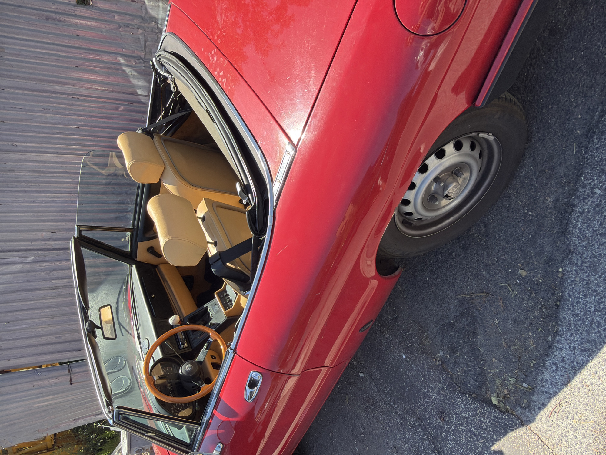 A red convertible Alfa Romeo spider is parked on an asphalt surface, possibly a street or parking lot, with a corrugated metal wall in the background. The car's top is down, revealing tan leather seats and a wooden steering wheel. The interior also shows a radio and a manual transmission shifter. The car appears to be in good condition, with minimal visible damage. The surrounding environment is likely outdoors, indicated by the natural lighting and shadows. There are no people visible in the image. The overall impression is of a vintage sports car in an urban setting. The location in Rome, Italy is not discernable from the visual evidence in the image.