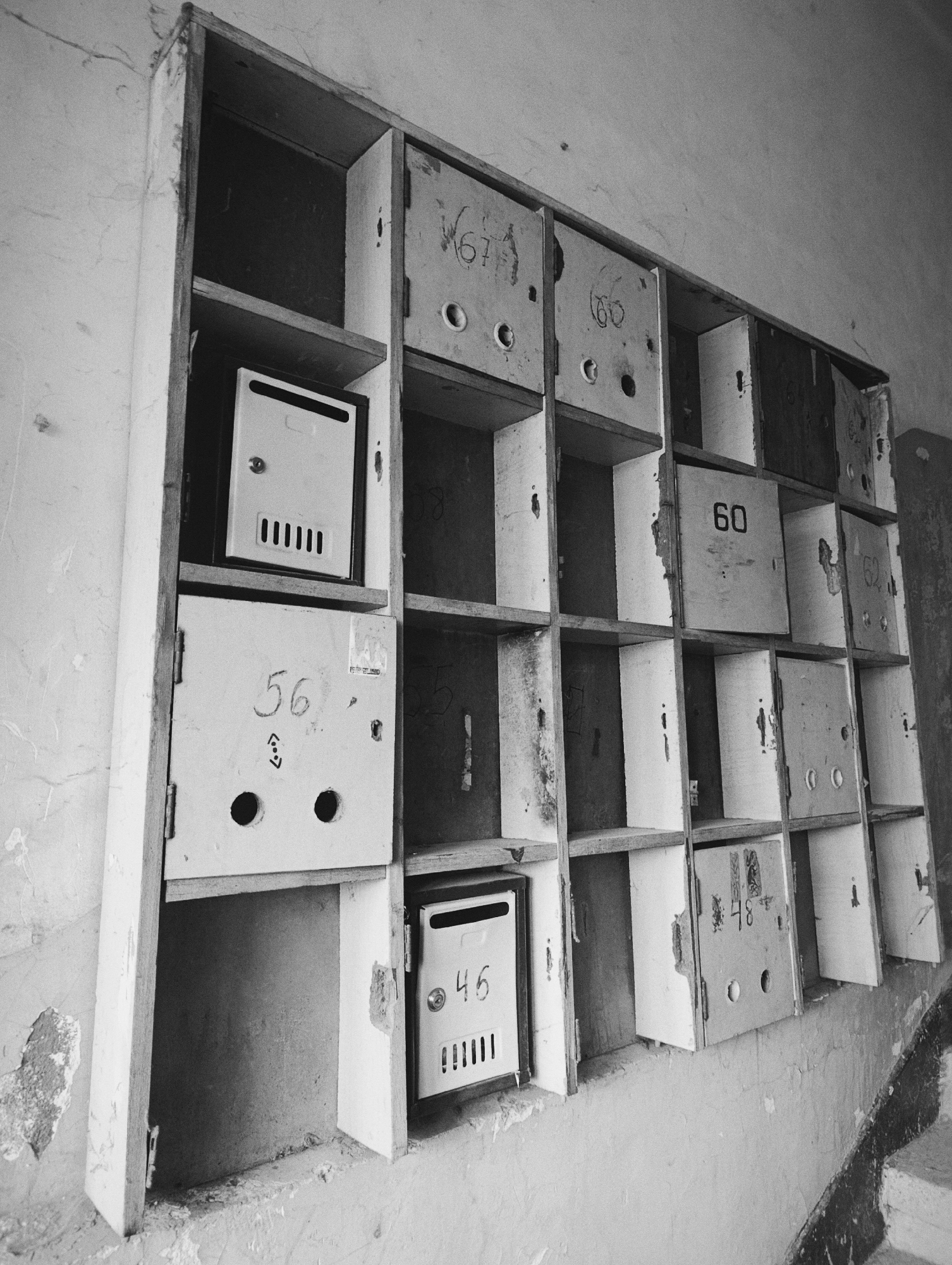 A black and white, low-angle shot shows a weathered wooden shelf containing mailboxes attached to a wall. The shelf is divided into several rectangular cubicles, each likely intended for a mailbox. Most of the mailboxes are missing or damaged, revealing empty space within the cubicles. Some remaining mailboxes are painted white, with visible numbers and holes. The paint on the mailboxes and surrounding shelf is peeling. A small metal mailbox is visible in one of the cubicles. The wall shows signs of decay, with peeling paint and cracks. The scene appears to be in a state of disrepair and neglect. The location is Kardzhali, Bulgaria.