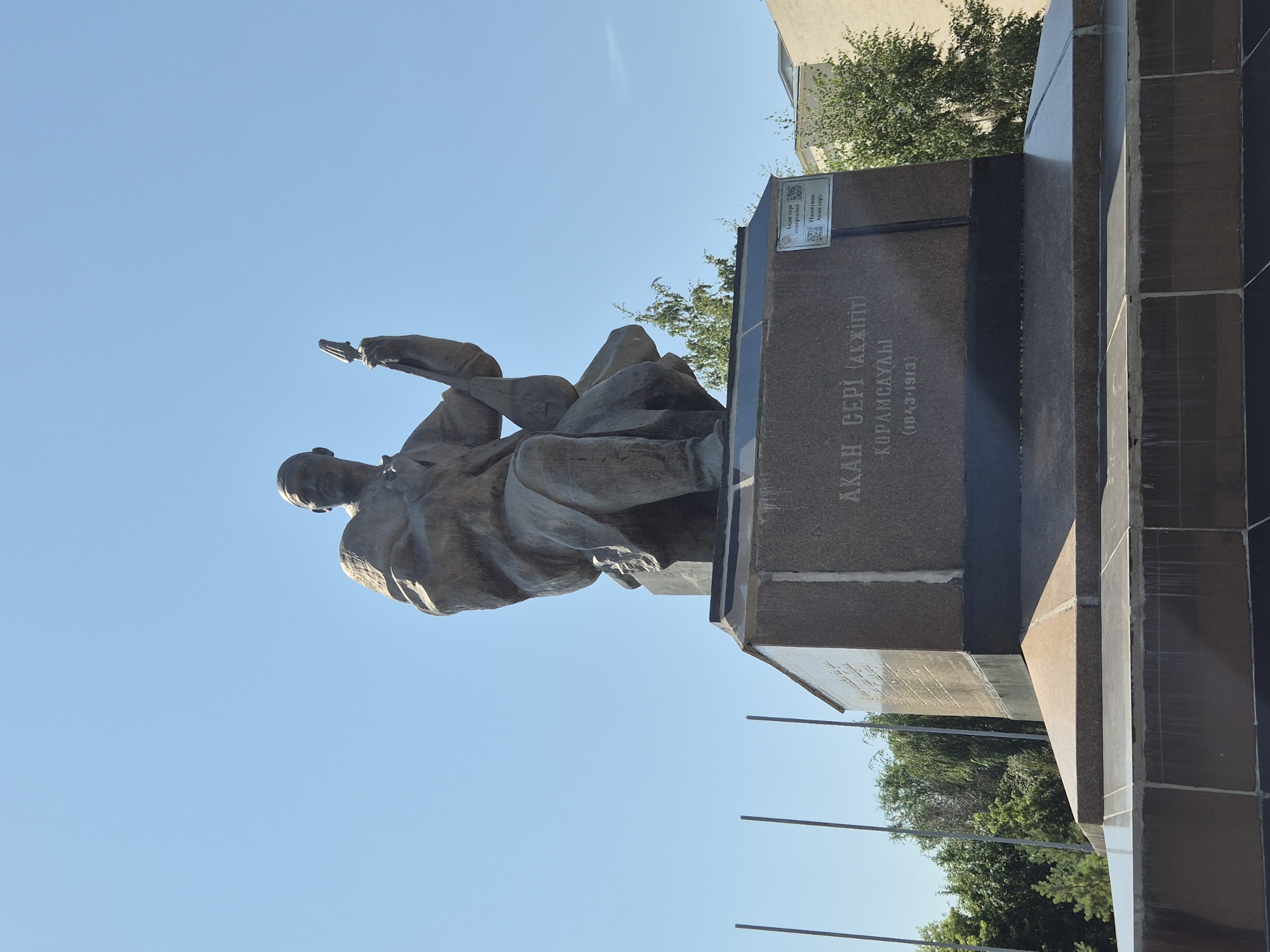 The image shows a statue of a man holding a dombra, a traditional Kazakh stringed instrument. The statue is made of bronze and is mounted on a stone pedestal. The pedestal has an inscription that reads "Ақан Сері (Ақжігіт)" which translates to "Akan Seri (Akyigit)". Below this, it says "Қорамсаұлы" and "(1843-1913)". There is also a QR code on a plaque attached to the pedestal. The statue is located in the city of Kokshetau, Kazakhstan, as indicated by the context. The background shows a clear blue sky, trees, and buildings. The overall scene appears to be outdoors, possibly in a park or public square.