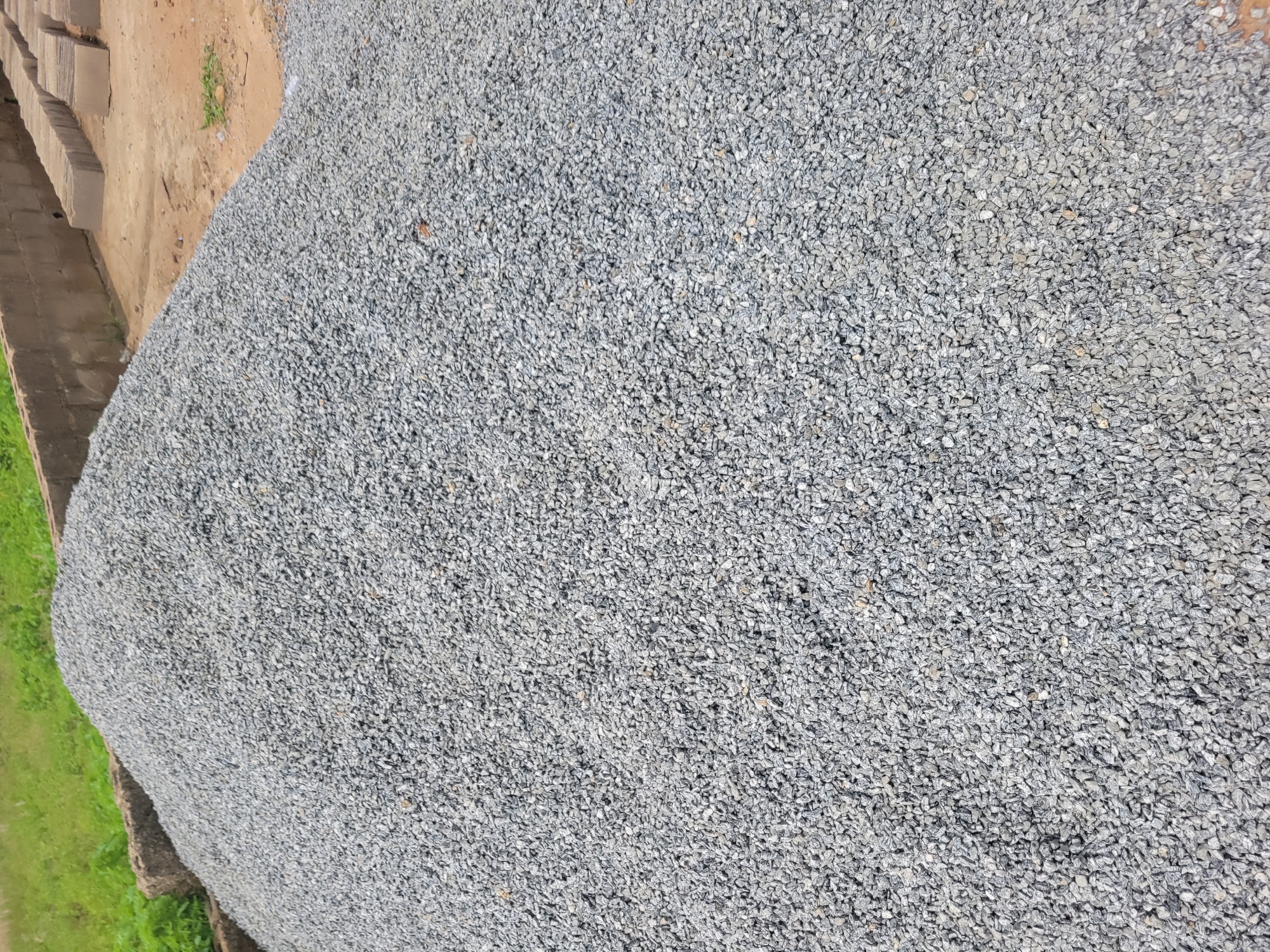 A pile of grey gravel is featured in the foreground of this image. The gravel is composed of small, angular stones with some variation in color from light grey to darker grey. The pile forms a large mound that dominates the center and right portions of the frame.
In the background, to the left, a patch of green grass and vegetation is visible. Behind the gravel pile and to the right, a low wall constructed of concrete blocks is present. Adjacent to the wall, stacked rows of rectangular concrete blocks, possibly bricks or pavers, are arranged on a sandy, reddish-brown surface. The overall environment appears to be an outdoor construction or material storage area. There are no people visible in the image.