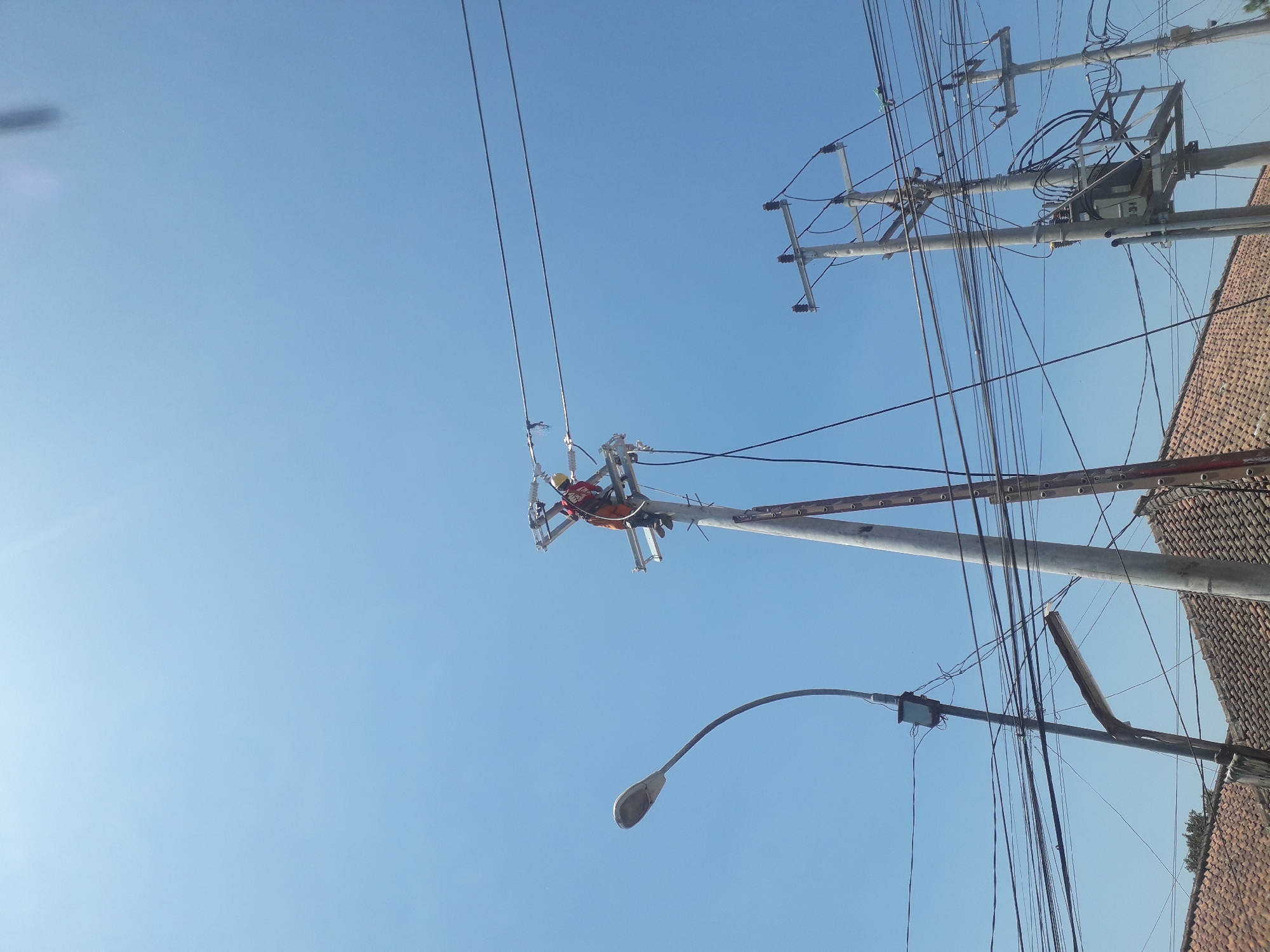 In Cankuang, Indonesia, a utility worker wearing an orange harness and a hard hat is actively working on a utility pole. The worker is positioned on a platform attached to the pole, attending to overhead electrical wires. The scene is dominated by a clear blue sky, with several utility poles and a streetlamp visible. Below the utility lines, the roofs of several houses are apparent. The overall image captures a moment of infrastructure maintenance against a bright, sunny backdrop.