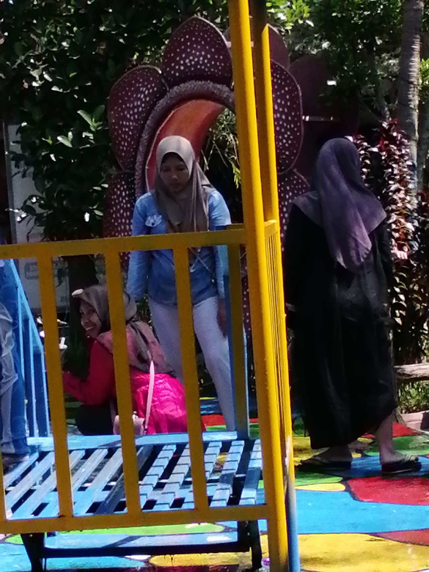 The image shows an outdoor scene, likely in a park or amusement area, with three individuals visible. In the foreground, a bright yellow metal railing runs horizontally and vertically, partially obscuring the view. Behind the railing, on a blue and red painted surface, a young person wearing a pink hijab and a bright pink garment is sitting. To their left, another person, also wearing a hijab and a light blue denim jacket over white pants, stands. Further to the right, a third person, wearing a dark outfit and a purple hijab, stands facing away from the camera. In the background, a large, stylized representation of a Rafflesia flower is visible, along with lush green foliage and a palm tree. The ground is painted with various bright colors, suggesting it may be a play area or a themed attraction. This scene is located in the city of Tegalwangi, Indonesia.