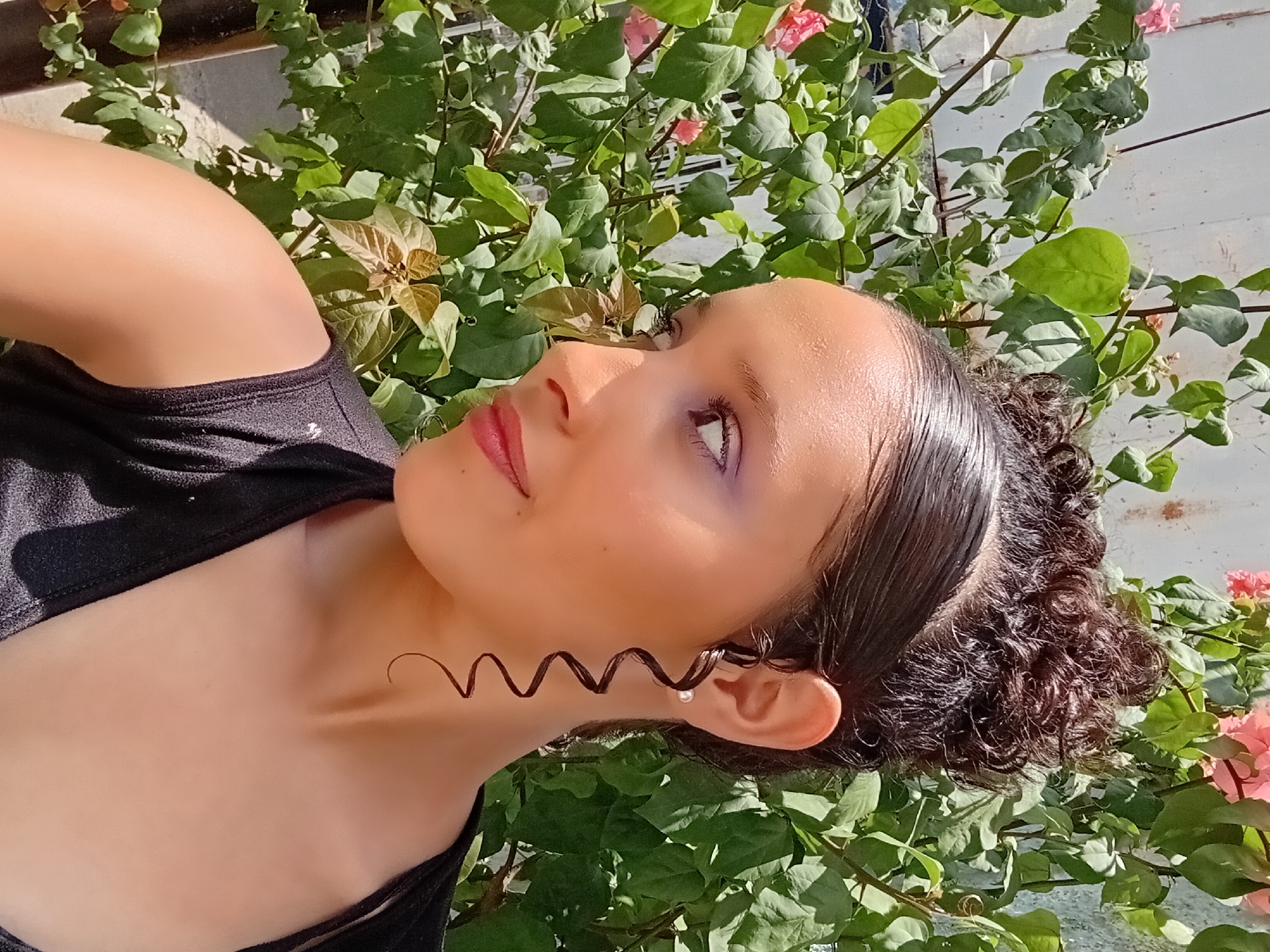 A medium close-up shot, rotated to show a person in a vertical orientation, captures a young individual posing outdoors amidst lush greenery. The main subject, appearing to be a female, is positioned in the upper right quadrant of the frame, looking upwards and slightly to her left, with a serene and gentle expression. Her head is tilted, and her profile is visible. She has dark, shiny, curly hair, pulled back, with a distinctive, well-defined spiral curl hanging forward near her ear, alongside a small, light-colored earring, possibly a pearl. Her skin appears smooth and is highlighted by bright sunlight, giving it a somewhat dewy or glossy appearance. She wears a dark-colored top, possibly black or dark gray, visible around her shoulder and neck area.
The person's face features soft makeup, including pink lipstick and subtle purple eyeshadow. Her eyes are open, looking towards the top left of the frame. The background is dominated by vibrant green leaves of climbing plants, interspersed with small clusters of bright pink flowers, likely bougainvillea, suggesting a garden or an outdoor setting like a balcony in a warm climate. A white or light-colored wall is visible through the foliage in the background. The bright, direct sunlight indicates it is daytime, casting clear highlights on the subject's skin and hair, and creating varied textures within the foliage. The overall impression is one of natural beauty and tranquility in an outdoor, sunlit environment. There is no visible text in the image. The location is specified as Parroquia Ocumare, Venezuela.