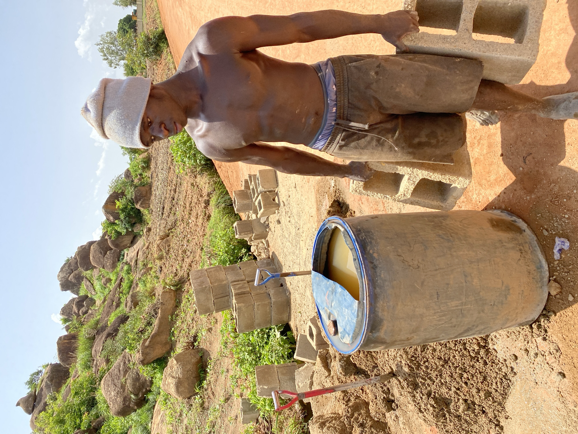 The image depicts a young Black male, shirtless and wearing a light-colored beanie and brown shorts, carrying two concrete blocks. He is standing on a dirt path in an outdoor setting. To his left, there is a large blue barrel filled with yellow liquid, with a shovel stuck in the ground next to it. In the background, stacks of concrete blocks are visible, along with a rocky hillside with sparse vegetation and trees under a bright, partly cloudy sky. The scene suggests a construction or manual labor site. The location is identified as Bukuru, Nigeria.
