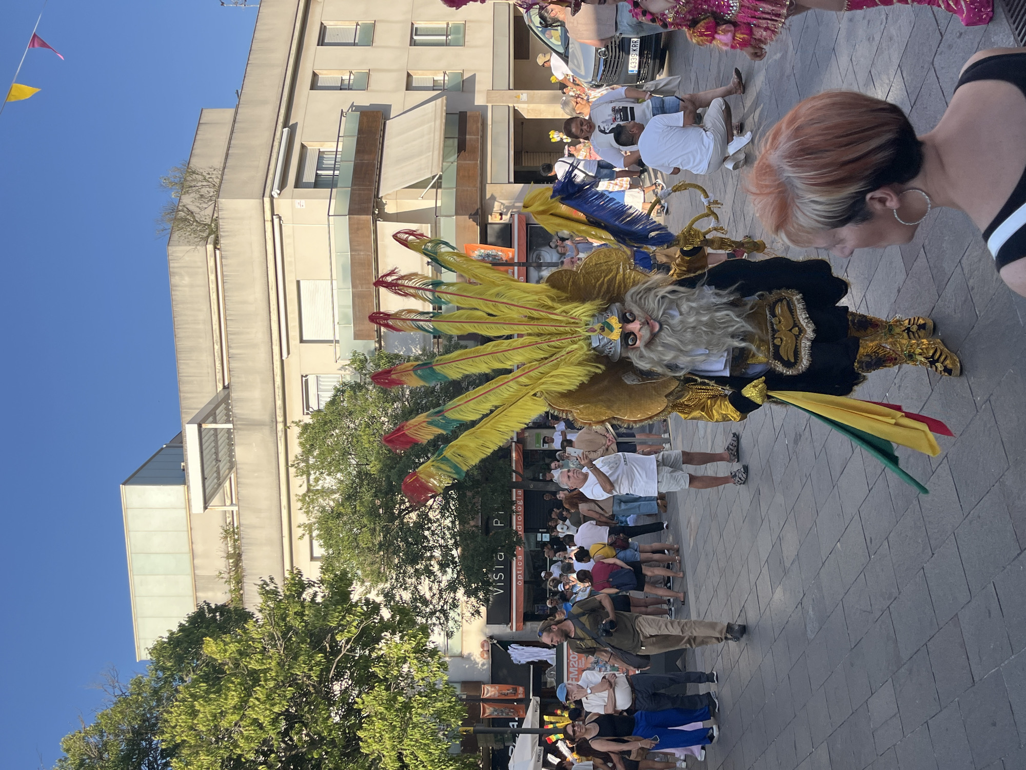 In a public square in Sant Cugat del Vallès, Spain, under a clear blue sky, a person in a large, elaborate costume is the central focus. The costume features a mask with a long white beard and intricate headdress adorned with yellow and red feathers. The person is also wearing a black and gold garment and ornate golden boots. They are holding a green, yellow, and red flag.
Several people are present in the square, some watching the costumed individual, others walking or standing. A white van with a license plate reading "4339 KRR" is visible on the right side of the frame. In the background, there is a multi-story building with balconies and shops. Trees are present on the left side of the square, and decorative flags are strung across the sky. The ground is paved with dark grey square tiles.