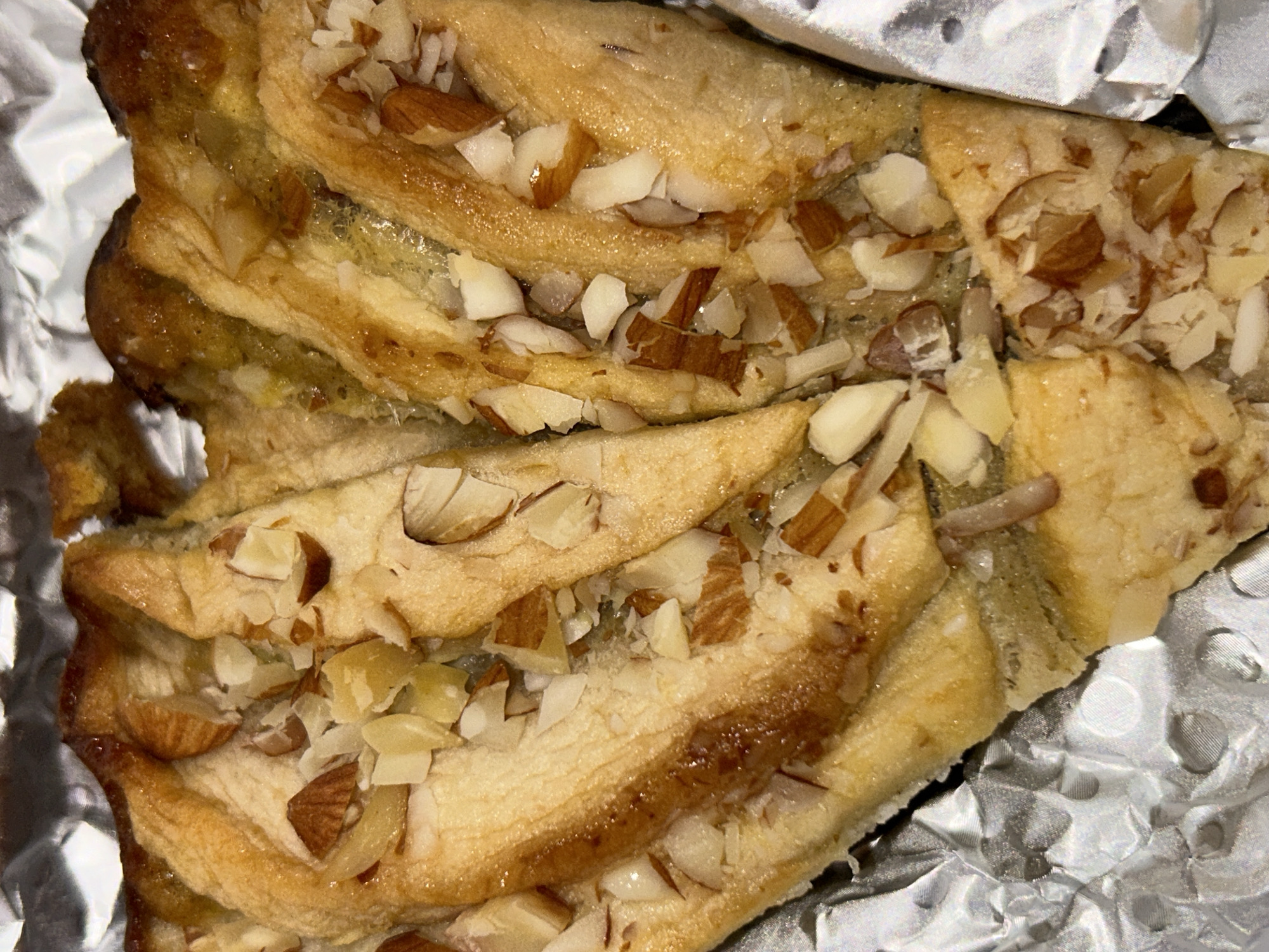 A close-up shot depicts a pastry, likely a sliced nut roll or strudel, resting on crumpled aluminum foil. The pastry has a layered, flaky texture with a golden-brown crust. Chopped almonds, both slivered and irregular pieces, are sprinkled liberally across the top surface. The foil provides a metallic, textured backdrop with highlights and shadows due to its crinkled surface. The scene does not provide explicit contextual information about the location being Sofia, Bulgaria.