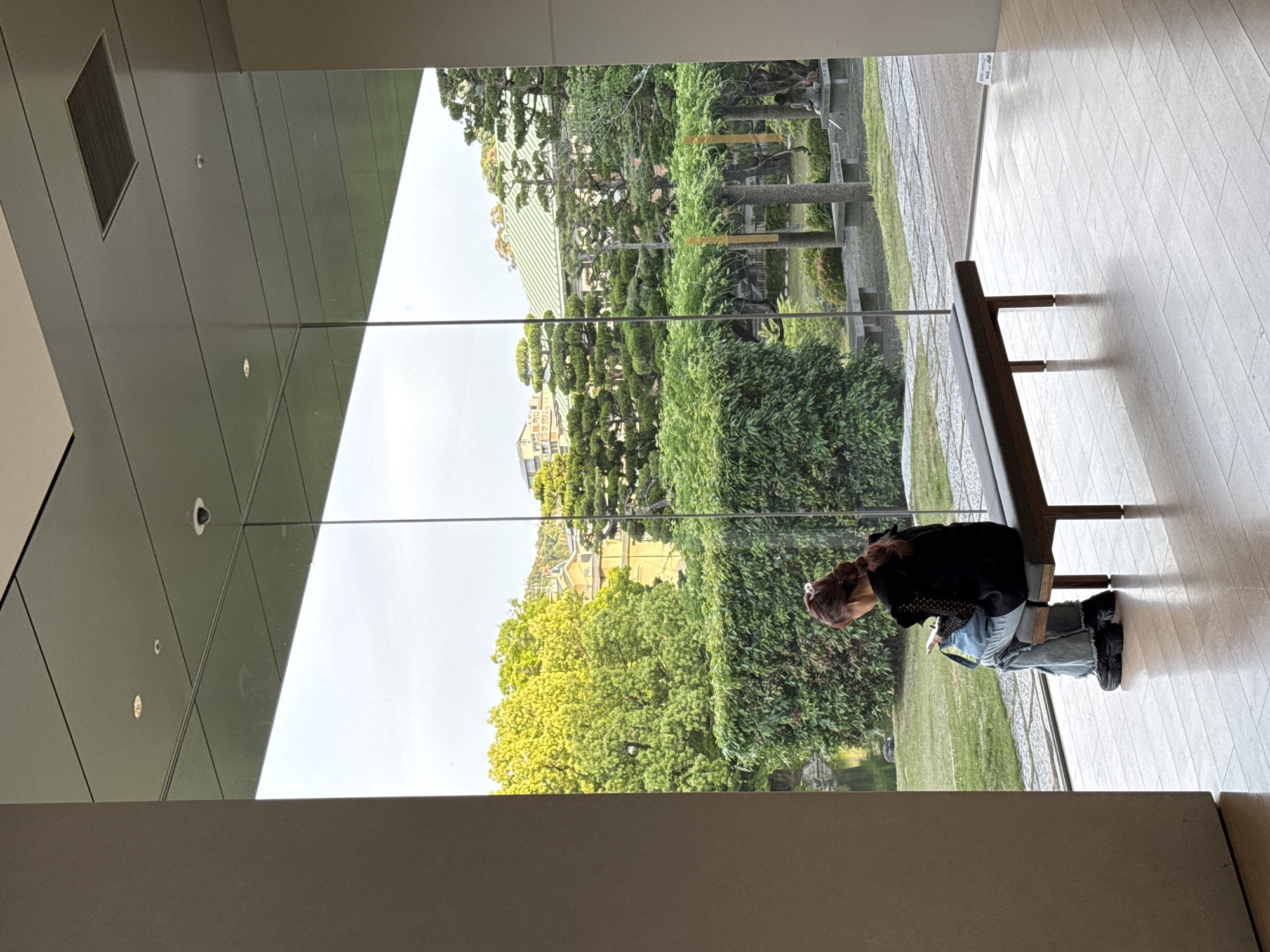 A medium shot taken indoors looks out through a large glass window onto a garden. A woman with dark hair, wearing a black jacket and jeans, is sitting on a wooden bench, facing away from the camera and towards the garden. The garden features manicured greenery, including trees and shrubs, and appears to be part of a larger, landscaped area. In the background, a light-colored building can be seen through the foliage.
The interior space is modern with grey walls and tiles on the floor. The perspective suggests the photo was taken from an elevated position within the building.
Based on the architecture and garden style, the location is likely in Kyoto, Japan, possibly within a cultural or historical site, like the Heian Shrine or Nijo Castle, known for their traditional Japanese gardens.