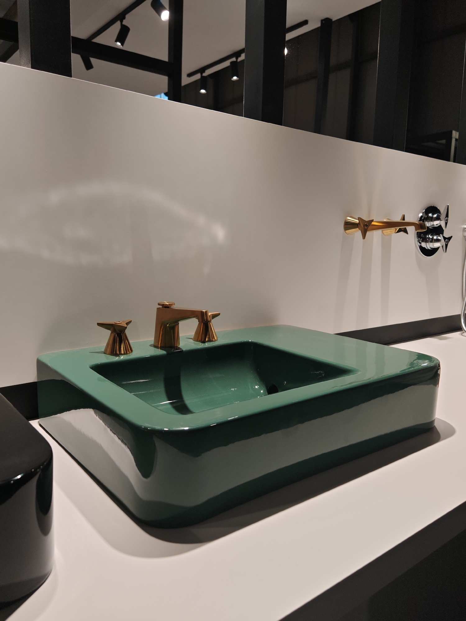 This is a vertical, eye-level shot showcasing a modern bathroom fixture display, likely in a showroom setting. The central subject is a rectangular, countertop-mounted sink in a vibrant, glossy deep green finish, featuring an integrated basin. Three brass-colored fixtures with distinctive, angular handles are installed on the back rim of the sink: a central faucet flanked by two smaller control handles.
The sink is set against a clean white wall and rests on a white countertop. To the right, mounted on the white wall, is another brass-colored faucet with a similar star-shaped handle design. Next to this wall-mounted faucet, a chrome fixture with a flexible hose, possibly a hand shower, is visible.
The background reveals the interior architecture, including black structural beams and track lighting with multiple spotlights on the ceiling, indicating an artificially lit indoor environment. Reflections are visible on the glossy surfaces of the green sink, the brass fixtures, and a dark, glossy, rectangular object partially visible in the bottom left foreground. No specific activity is taking place, as it's a static display. There is no visible text in the image.